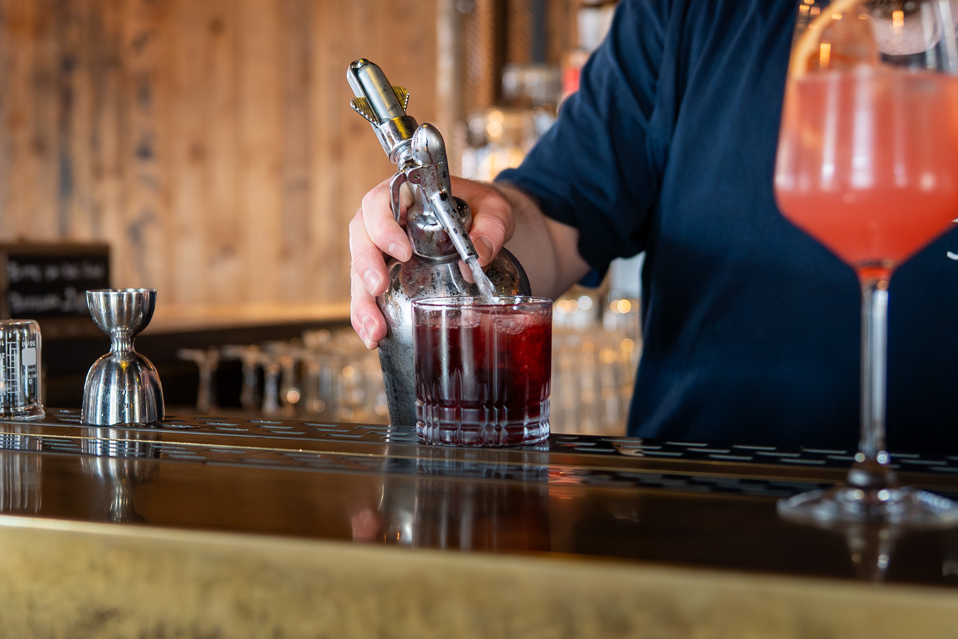 Zwei Gläser auf einer Theke, eines ist mit einer rosafarbenen Flüssigkeit gefüllt, das andere mit einer dunkelroten. In ein Glas wird gerade Sodawasser hinzugefügt.