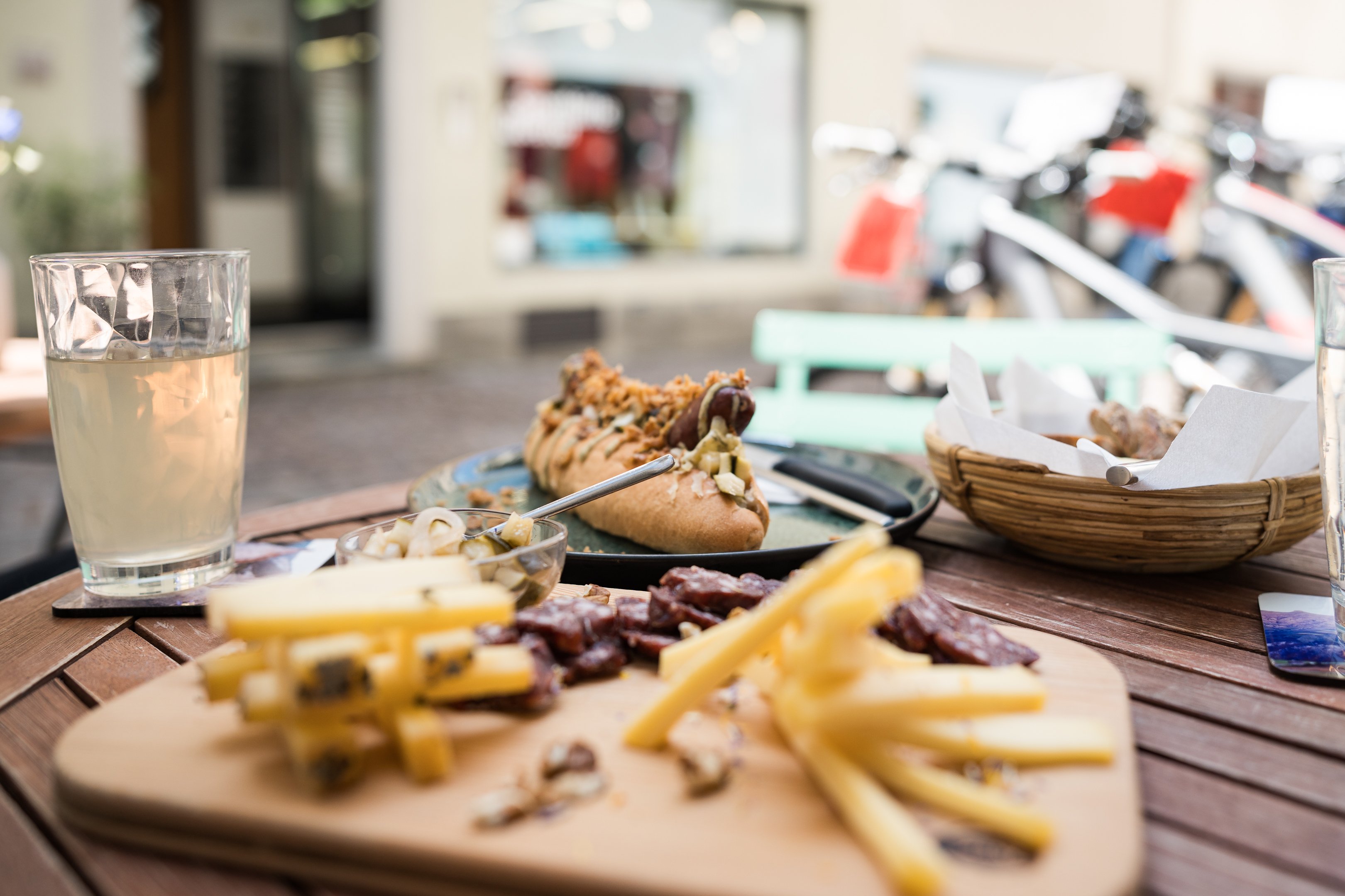 Un tavolo apparecchiato all’aperto con un hot dog, un tagliere di affettati e formaggi e un bicchiere di tè freddo.