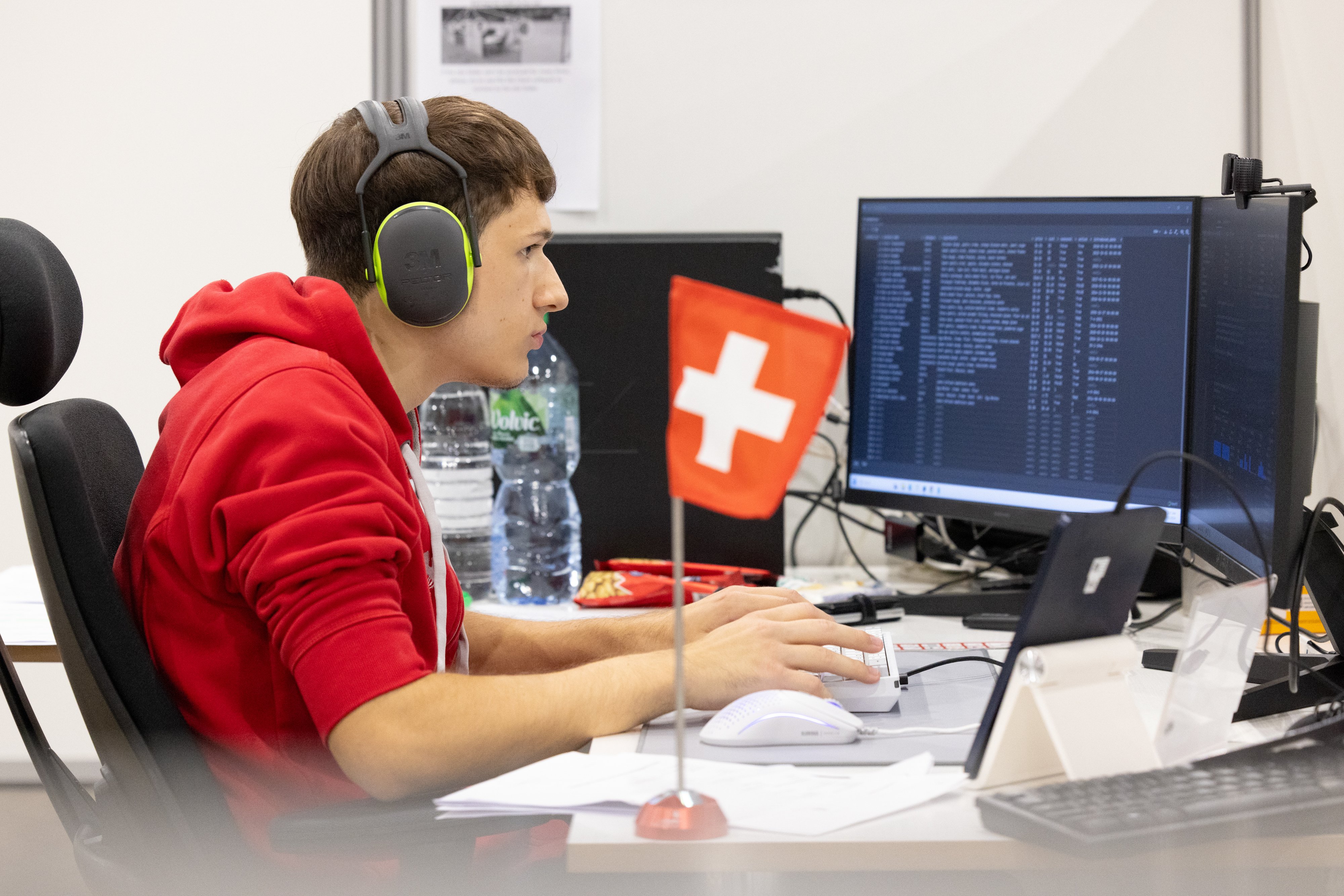 Besart est assis devant deux écrans et tape à la machine. A côté de lui se trouve un petit drapeau suisse.