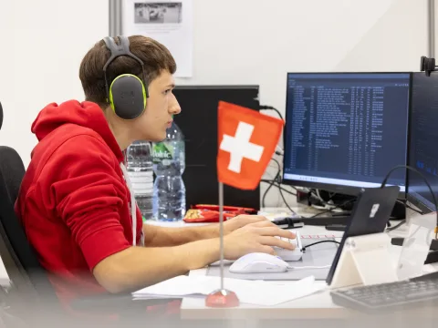 Besart est assis devant deux écrans et tape à la machine. A côté de lui se trouve un petit drapeau suisse.