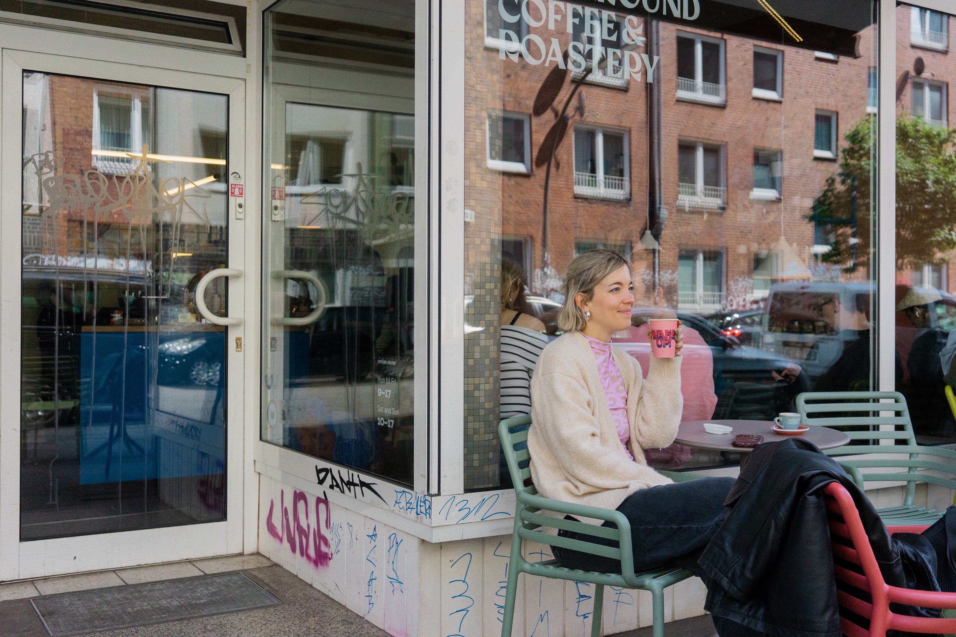 Eine Frau sitzt vor einem Kaffee und trinkt einen Kaffee während sich die Fassaden im Glas spiegeln.