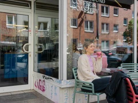 Eine Frau sitzt vor einem Kaffee und trinkt einen Kaffee während sich die Fassaden im Glas spiegeln.