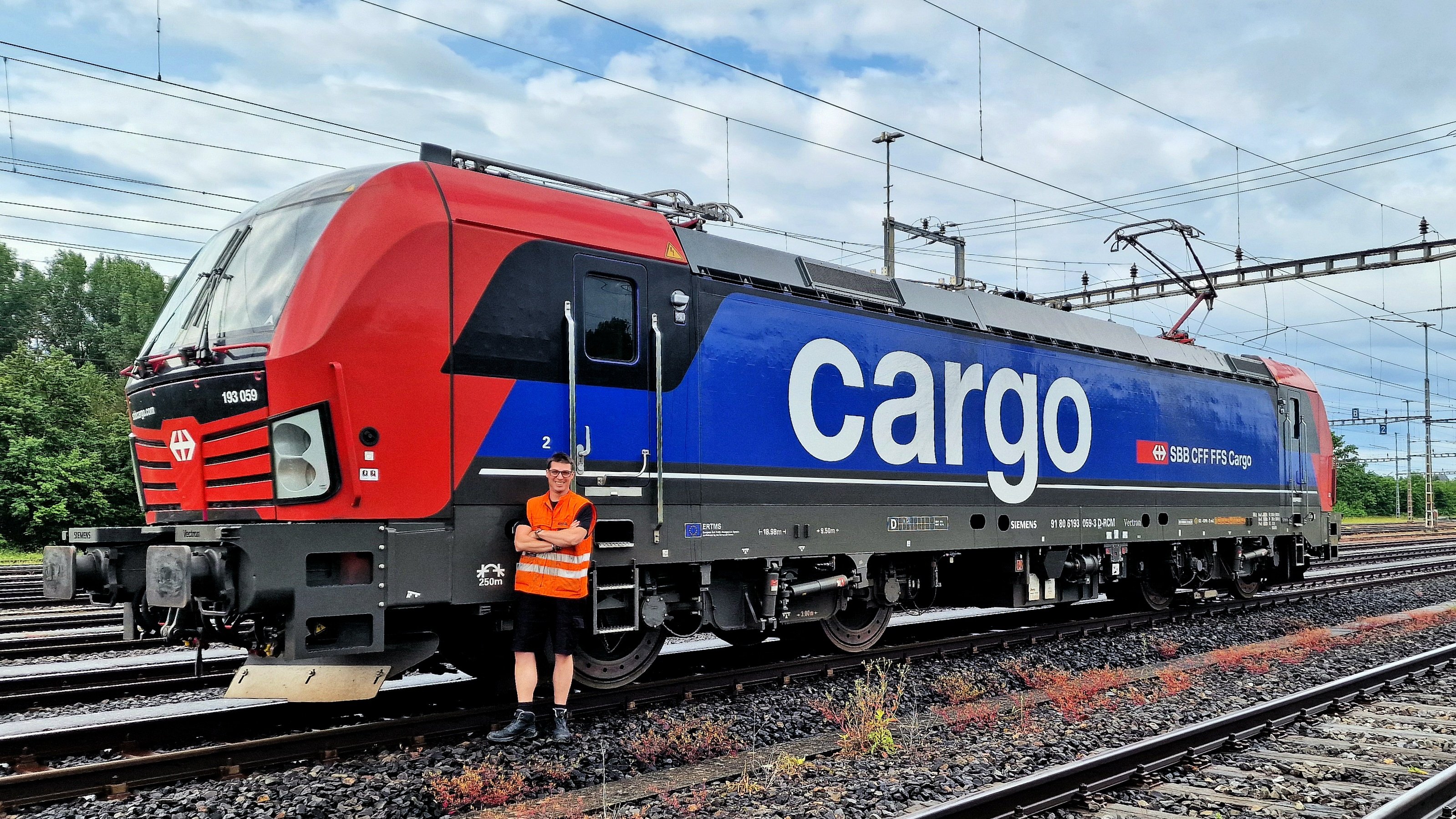Au dépôt de Lausanne Triage, le mécanicien Gilles Durgniat pose devant une locomotive de ligne Vectron. Il se tient debout, souriant et les bras croisés, à côté de l’échelle qui mène à la cabine de conduite. On constate que la locomotive est neuve.