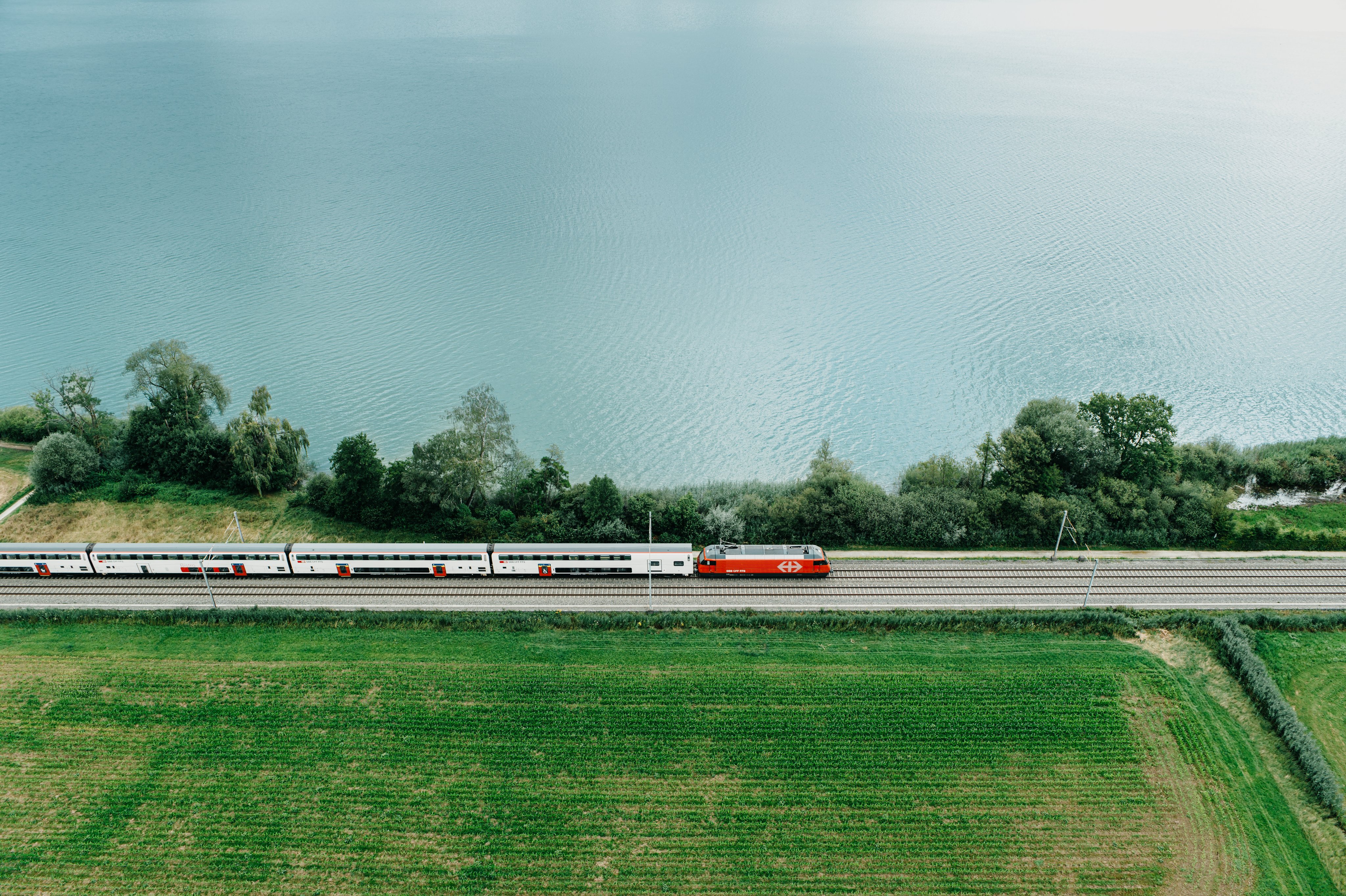 Eine rote Re 460 der SBB fährt zwischen einer Wiese und dem Genfersee entlang. Das Foto wurde mit einer Drohne von der Luft aus gemacht. Die Lokomotive zieht modernisierte IC 2000 Doppelstockwagen. 