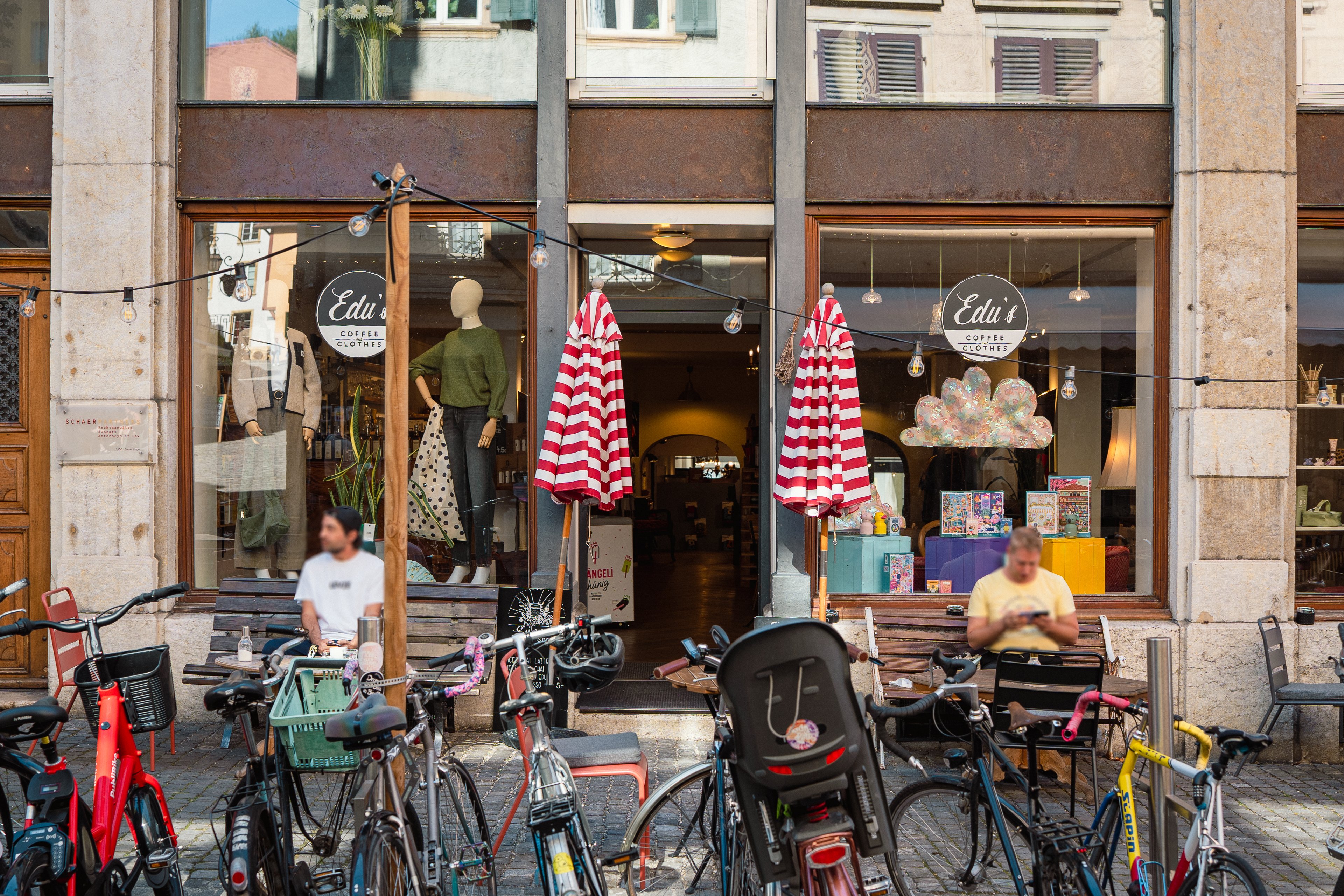 Davanti al negozio Edu’s nel centro storico di Bienne sono parcheggiate diverse biciclette. Accanto all’ingresso sono sedute due persone con delle bevande; sullo sfondo si vedono le vetrine del negozio.