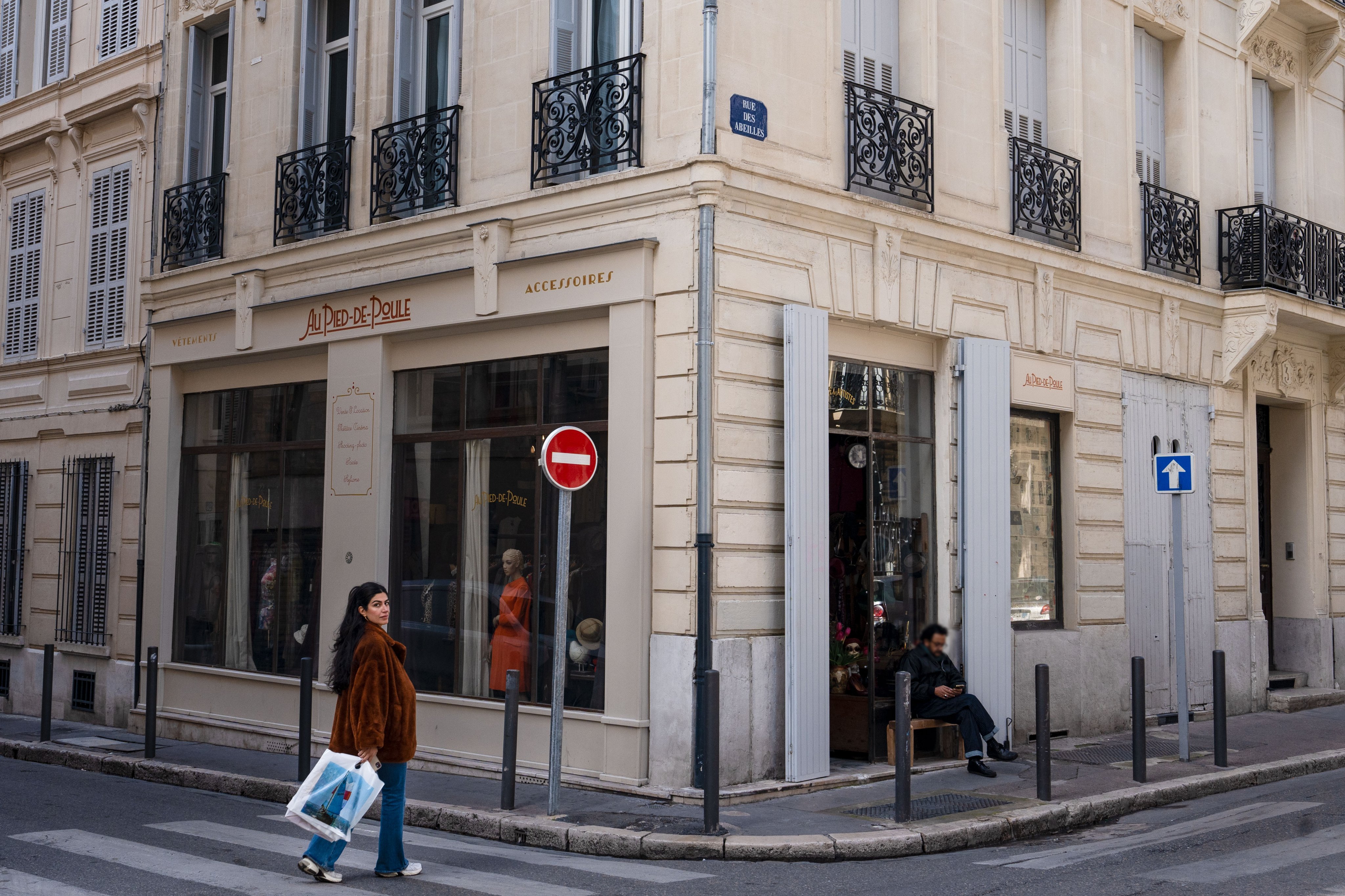 Une personne, à Marseille, passant devant la friperie «Au Pied de Poule» située au coin d’une rue, laissant entrevoir un grand nombre de vêtements à travers la vitrine.
