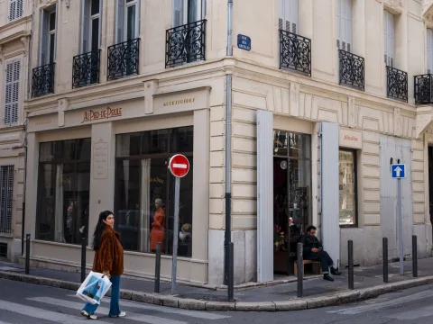 Une personne, à Marseille, passant devant la friperie «Au Pied de Poule» située au coin d’une rue, laissant entrevoir un grand nombre de vêtements à travers la vitrine.