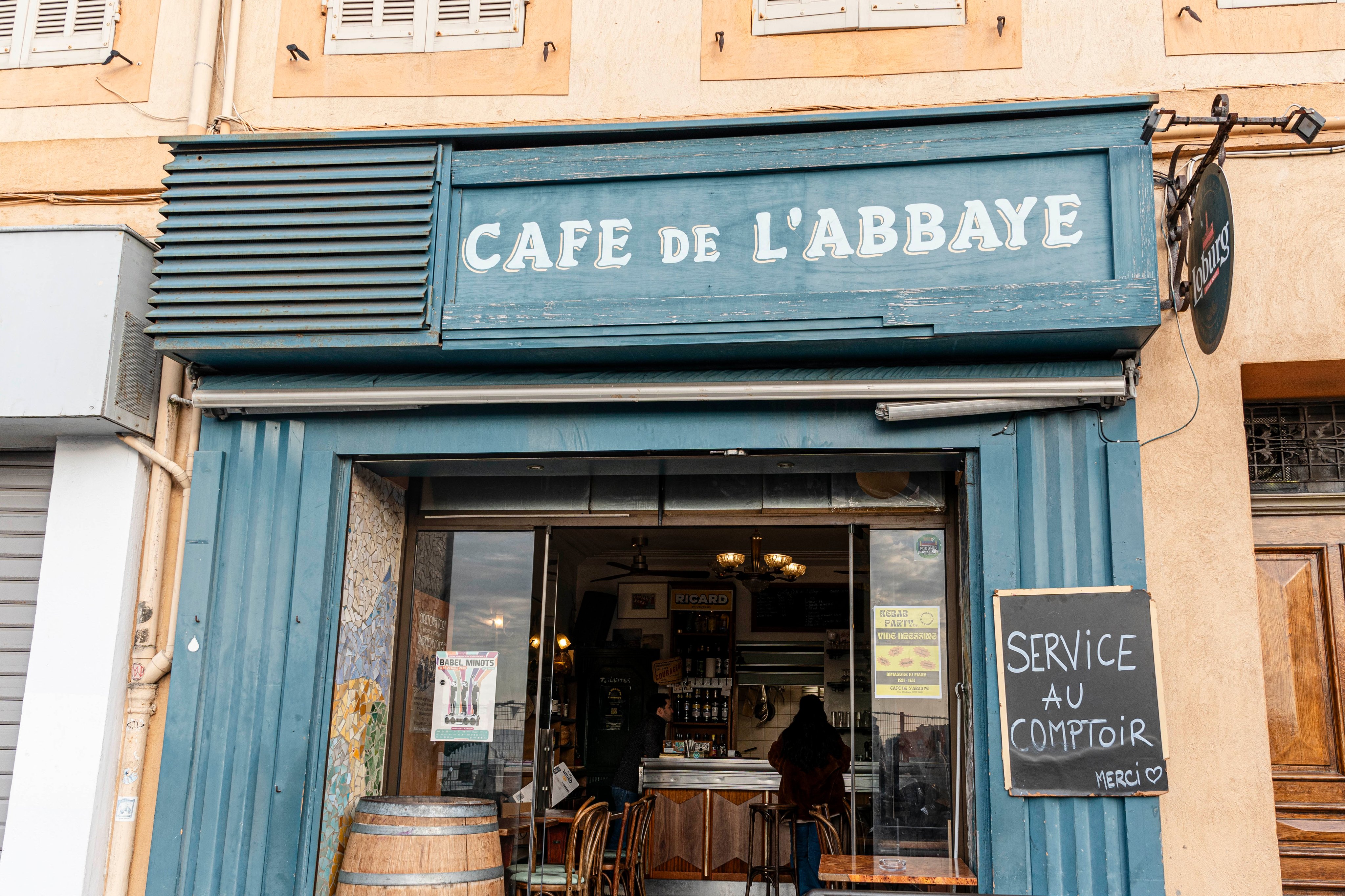 Vue de face du café de l’Abbaye à Marseille, avec sa traditionnelle marquise bleue et ses panneaux invitant à passer commande au comptoir.