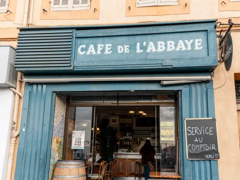 Vue de face du café de l’Abbaye à Marseille, avec sa traditionnelle marquise bleue et ses panneaux invitant à passer commande au comptoir.