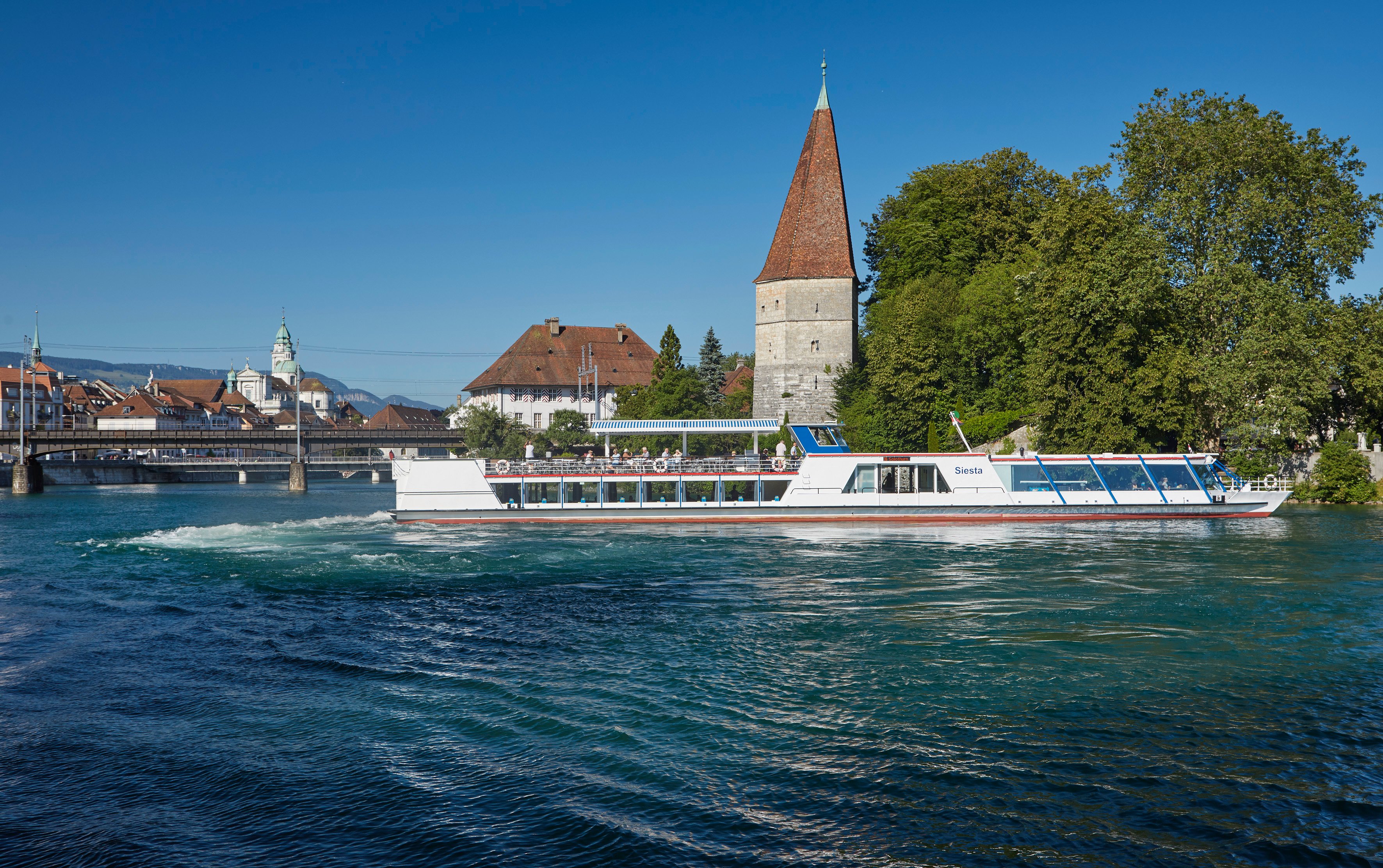 Ein Schiff auf einem Fluss, im Hintergrund ein Turm und Häuser.