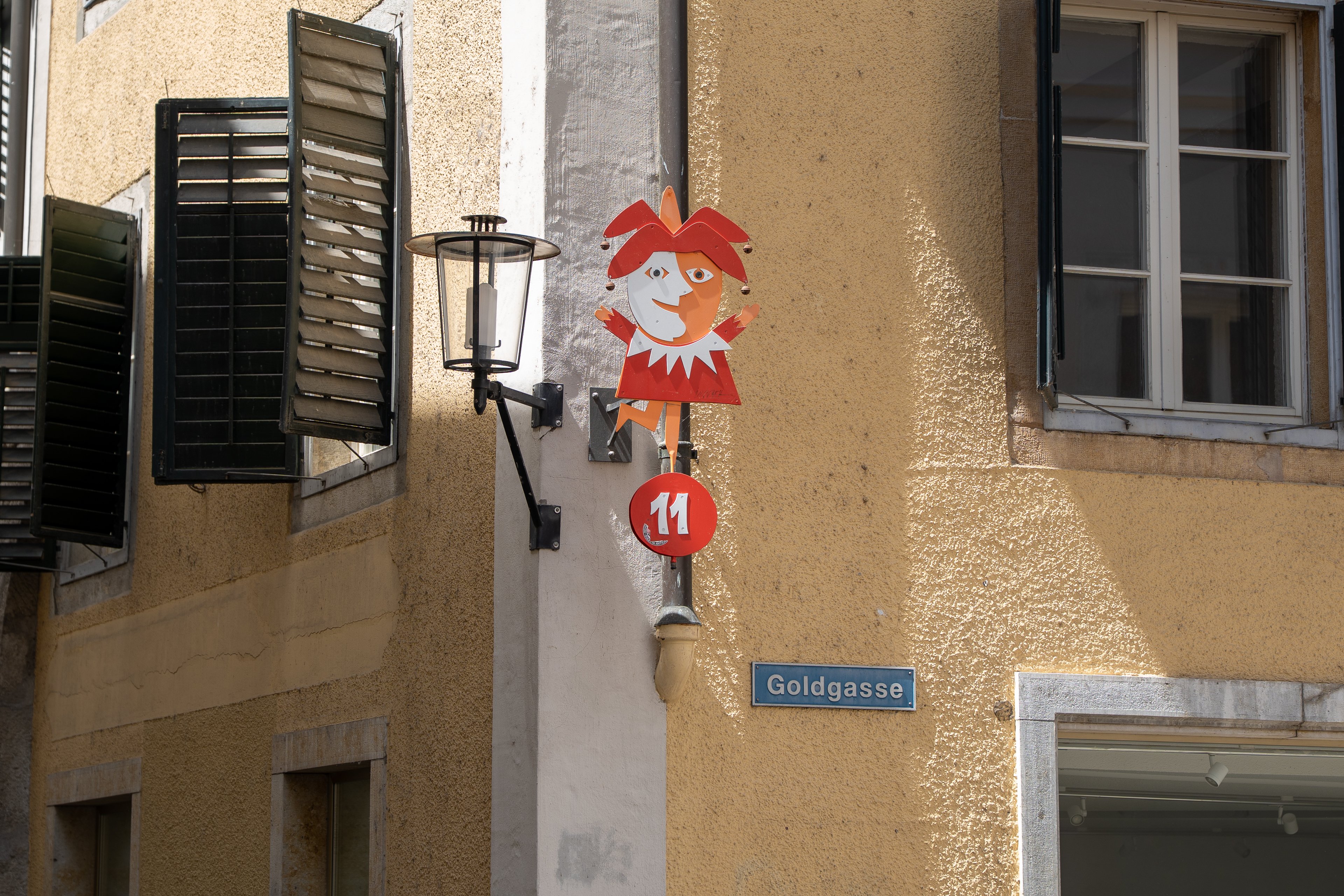 Hausfassade, an der ein Strassenschild, eine Laterne sowie eine Narrenfigur mit der Zahl elf angebracht ist.