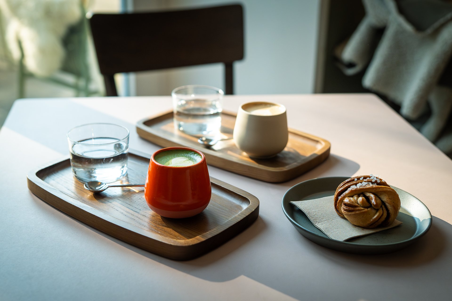 Auf einem Tisch stehen zwei Serviertabletts aus Holz mit je einer gefüllten Tasse und Wasserglas. Daneben ein Teller mit einem Gebäck.
