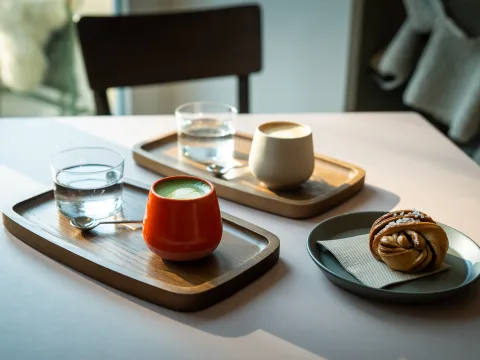 Auf einem Tisch stehen zwei Serviertabletts aus Holz mit je einer gefüllten Tasse und Wasserglas. Daneben ein Teller mit einem Gebäck.