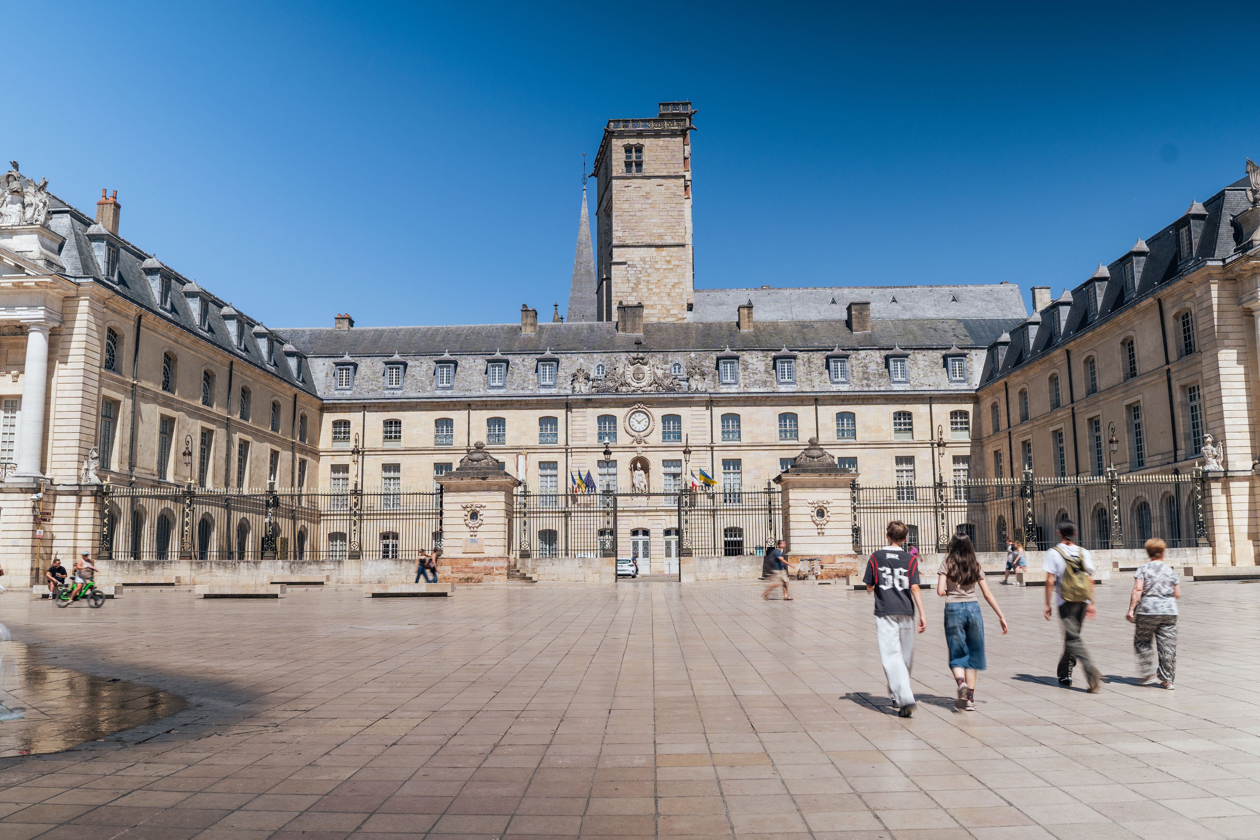 Un’ampia piazza con edifici disposti a U, alcuni passanti e una fontana. 