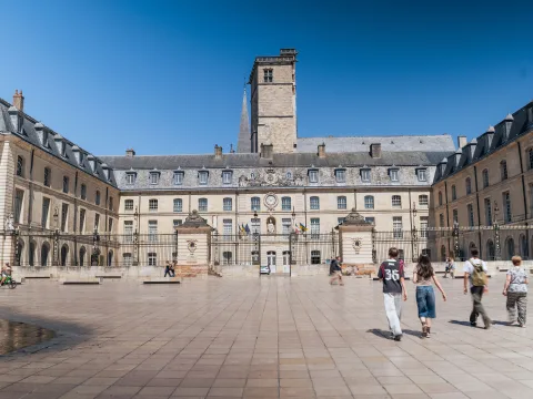 Un’ampia piazza con edifici disposti a U, alcuni passanti e una fontana.