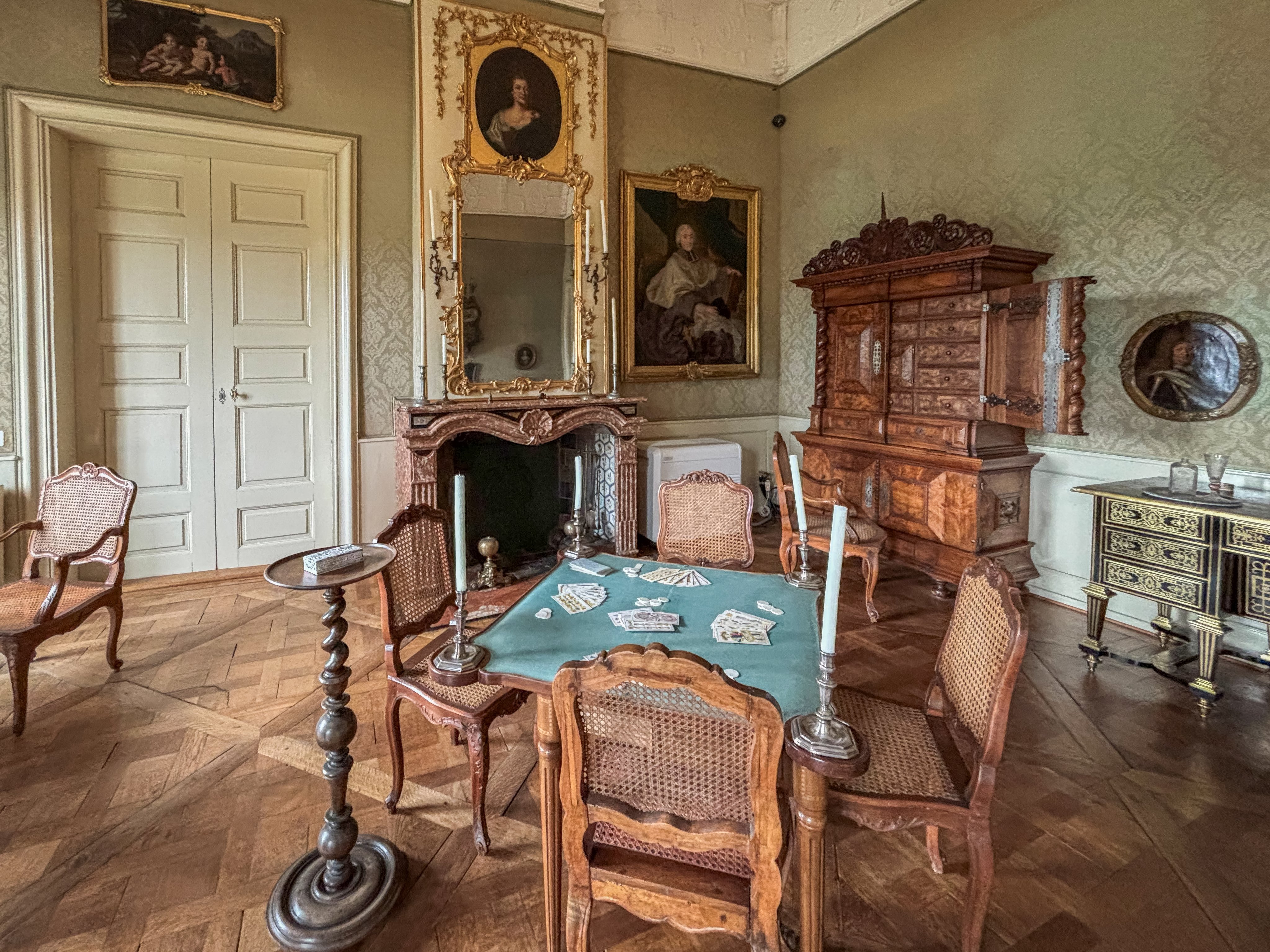 Ein Zimmer mit einem Tisch und vier Stühlen in der Mitte. Im Hintergrund gibt es ein Cheminée, und an einer Wand stehen eine Kommode sowie ein Sekretär.