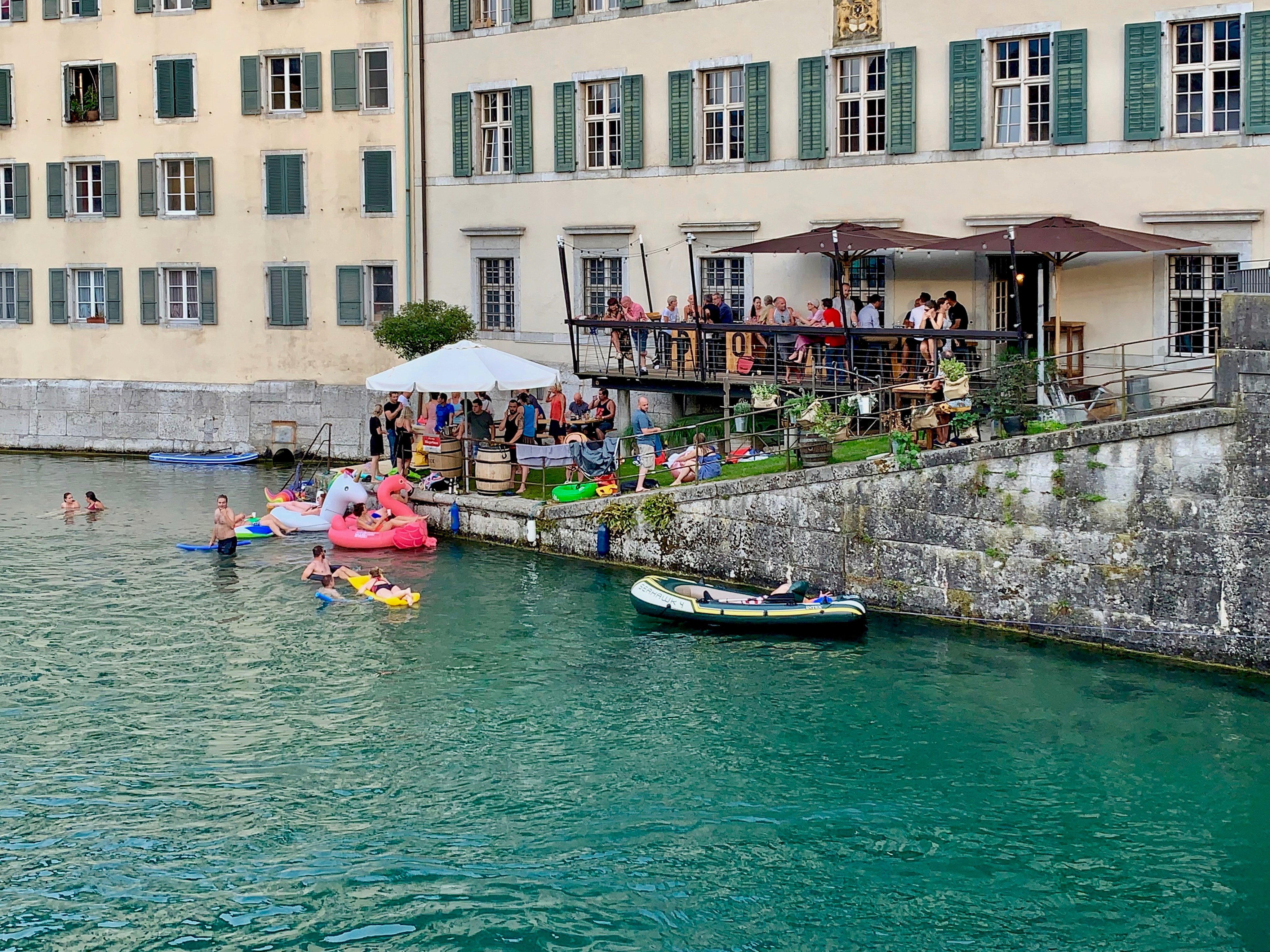 Ein Gebäude mit einer Terrasse und einer Rampe ins Wasser. Im Wasser schwimmen Menschen und Gummiboote.