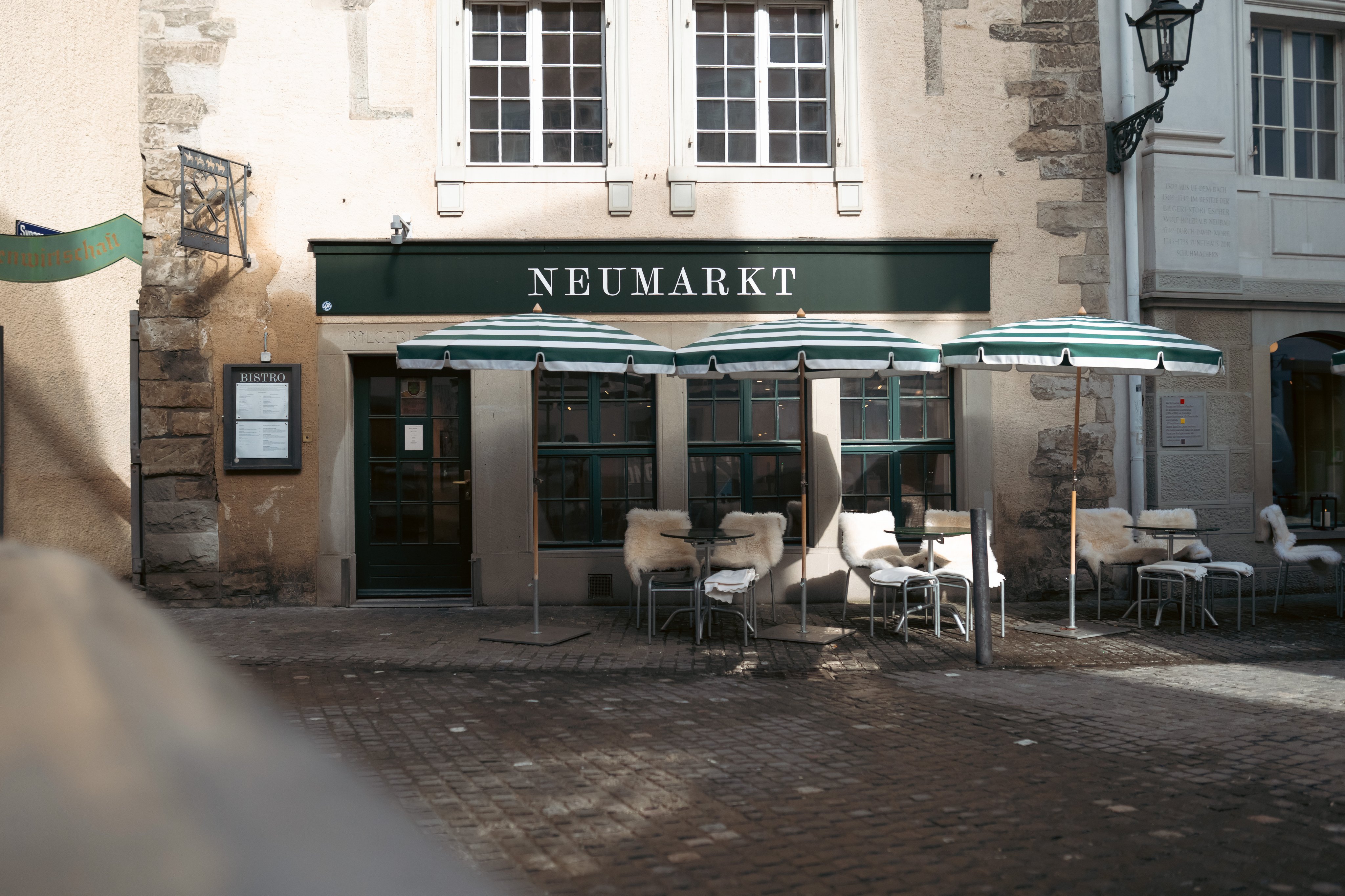 Un restaurant dont on voit le nom «Neumarkt» avec sa véranda, des sièges et des parasols.  