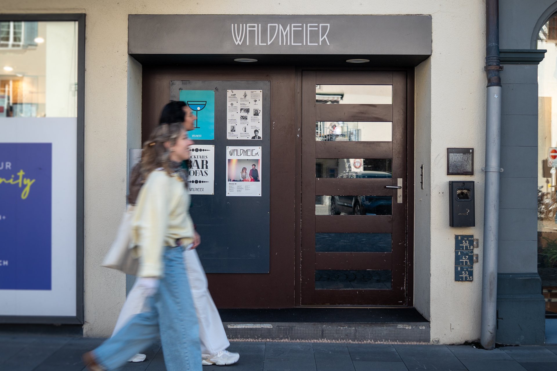 Zwei Frauen gehen an einer Bar vorbei. Über dem Eingang hängt ein Schild mit der Aufschrift Waldmeier.