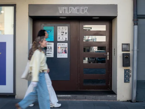 Zwei Frauen gehen an einer Bar vorbei. Über dem Eingang hängt ein Schild mit der Aufschrift Waldmeier.