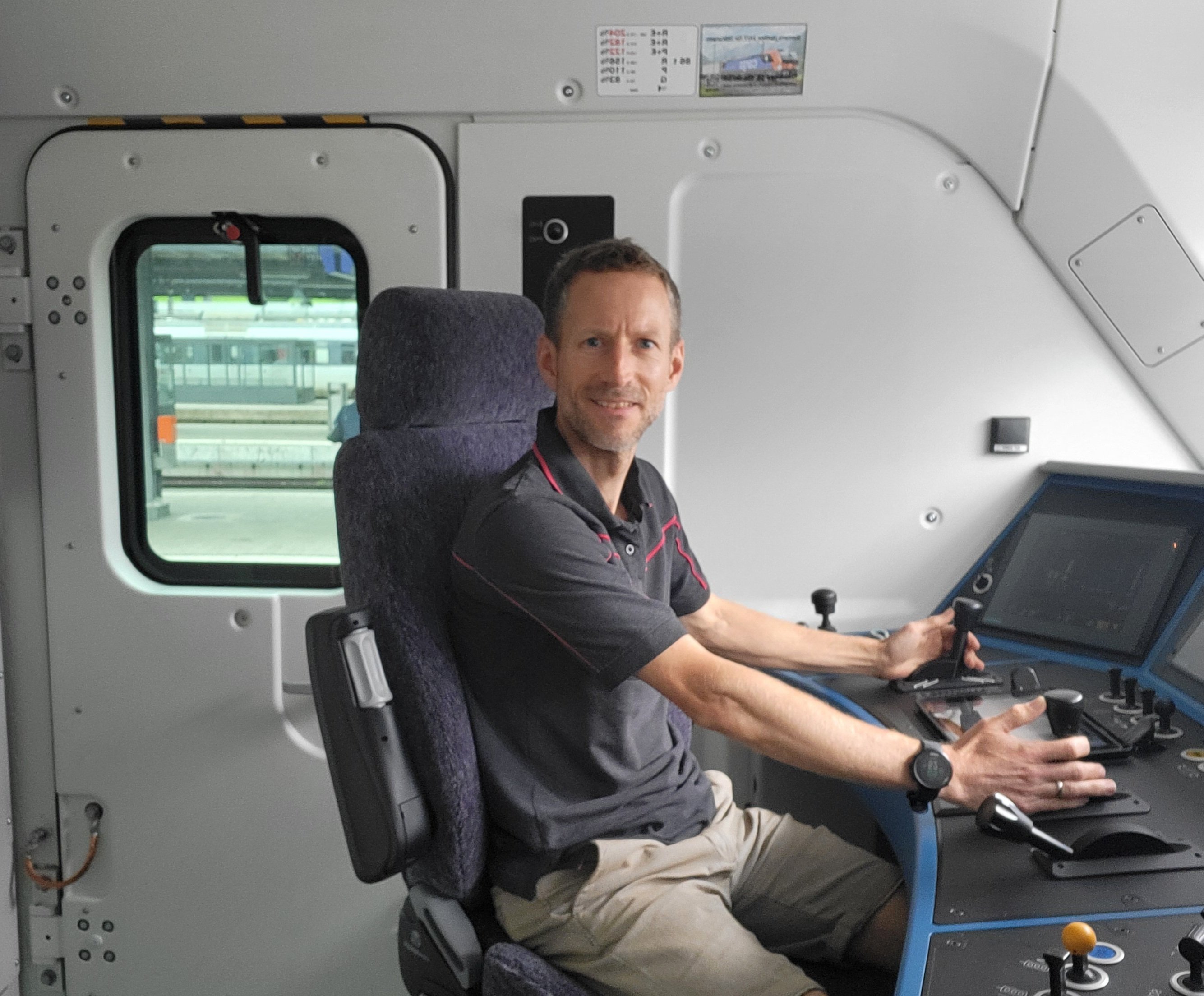Daniel Wüst, mécanicien de locomotive du dépôt de la RBL, est assis dans la cabine de conduite de la locomotive Vectron. Il regarde la caméra vers la droite en souriant aux mains posées sur les commandes de la locomotive. On reconnaît la fenêtre latérale de la cabine de conduite en arrière-plan.