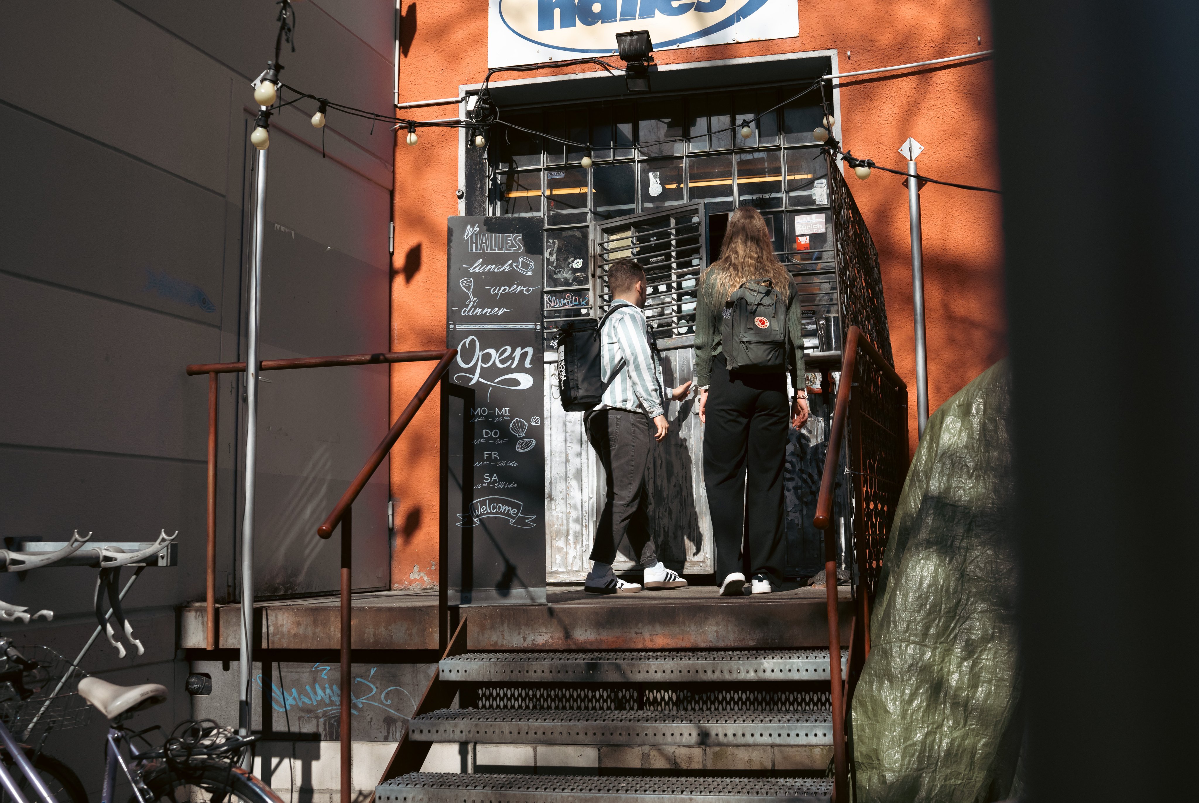 Deux personnes entrent dans un bâtiment rouge ressemblant à une usine. Au-dessus de l’entrée, il y a un panneau avec une inscription en bleu.  