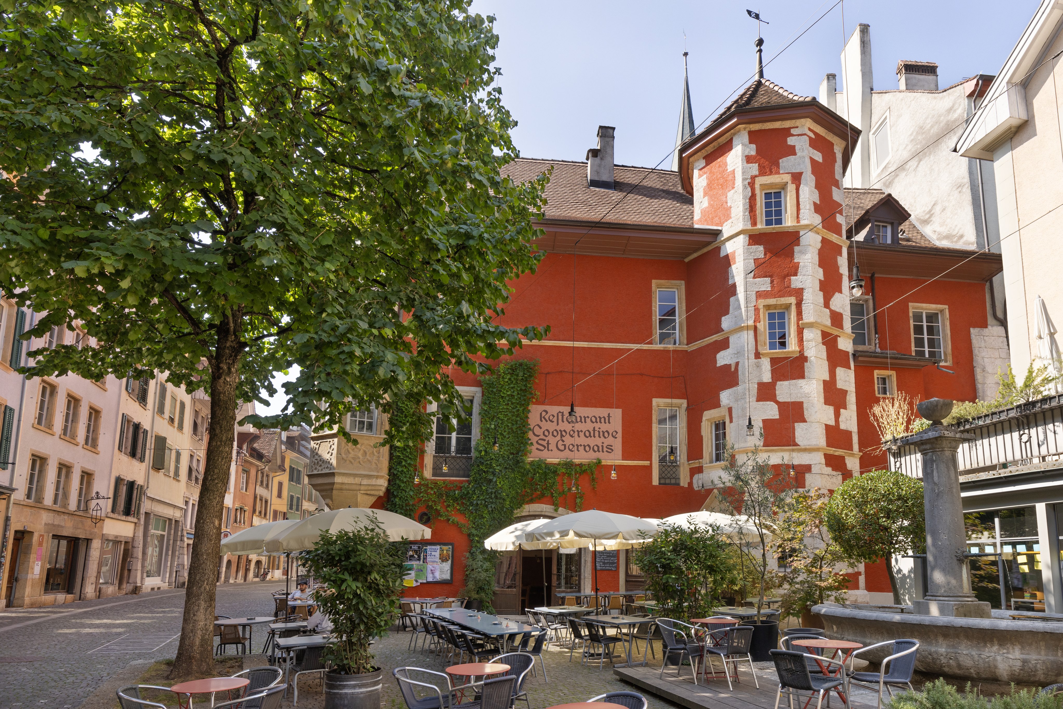 Piazza nel centro storico di Bienne davanti a un edificio, con tavoli e sedie. Diversi ombrelloni fanno ombra e, sul lato sinistro, un grande albero ripara dal sole una parte dei posti a sedere.