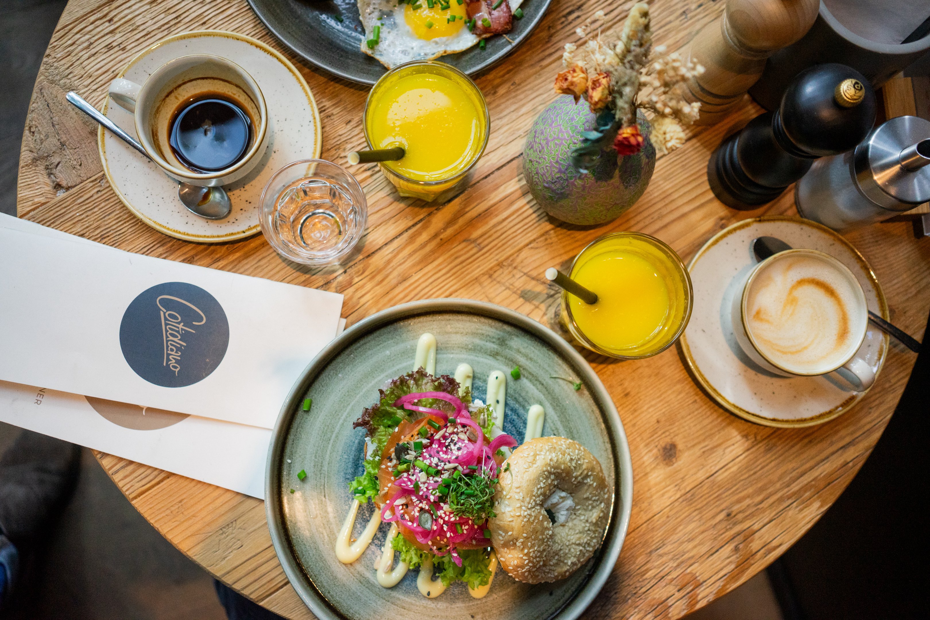 Vogelperspektive auf einen gedeckten Tisch mit Orangensaft, Kaffee und einem gut gefüllten Bagel.