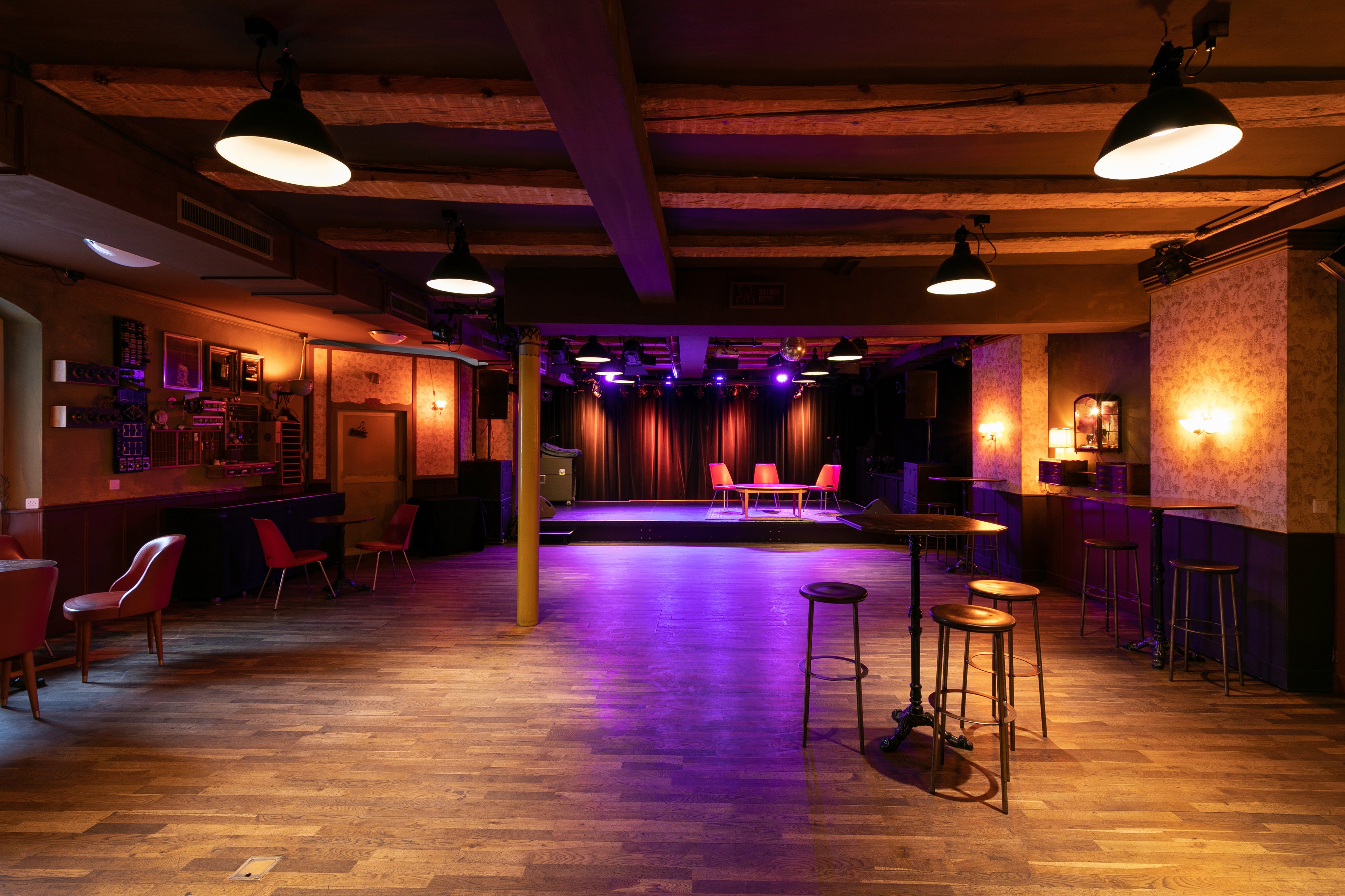 Interno del locale culturale «Le Singe», con pavimento in parquet, diversi tavoli e sedie e un piccolo palco sullo sfondo. L’ambiente è immerso in una suggestiva luce viola.