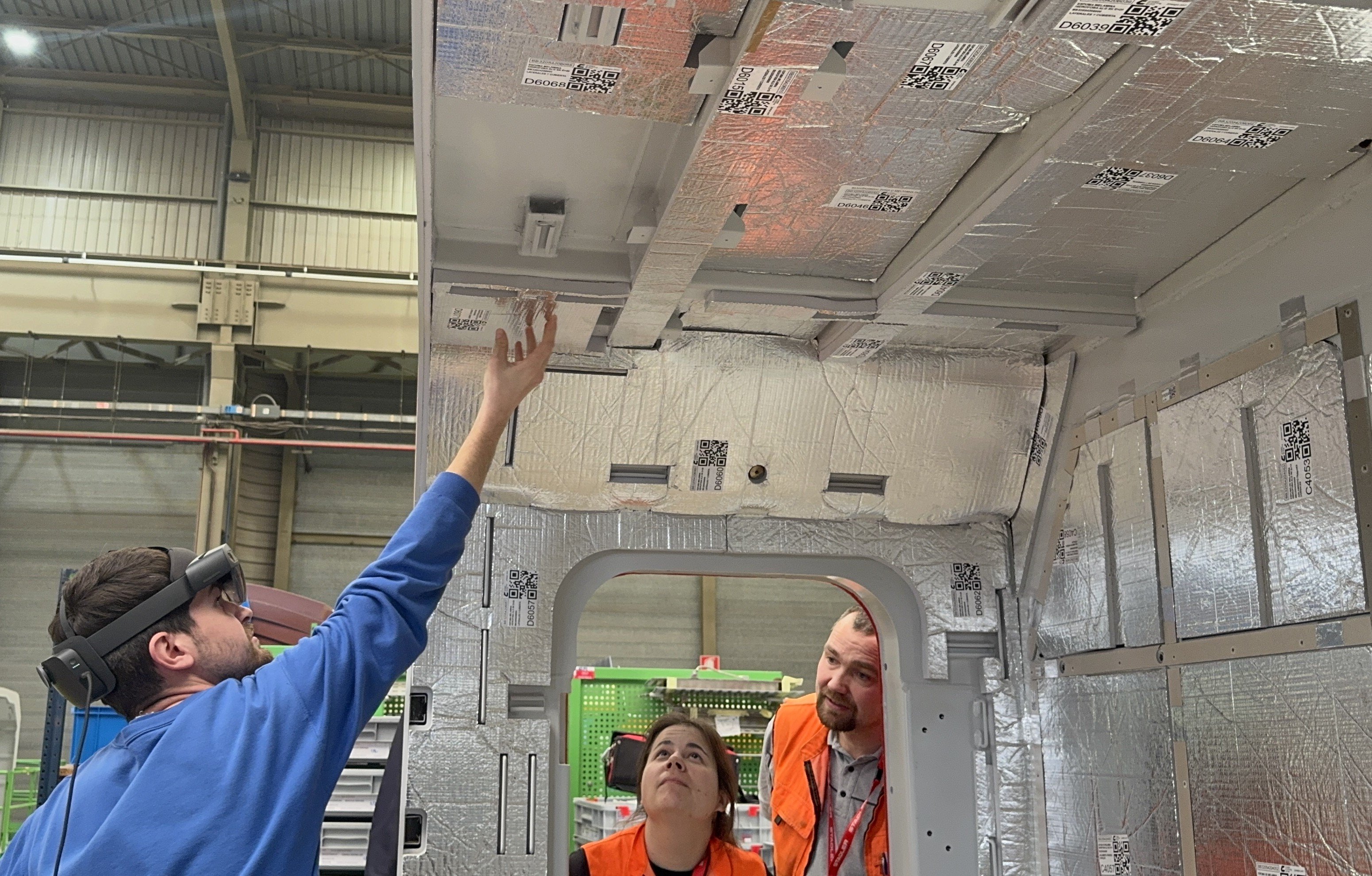 Un collaboratore della produzione posiziona del materiale isolante sul soffitto della cassa della locomotiva. Due collaboratori delle FFS osservano i lavori attraverso un finestrino della costruzione grezza di una locomotiva.