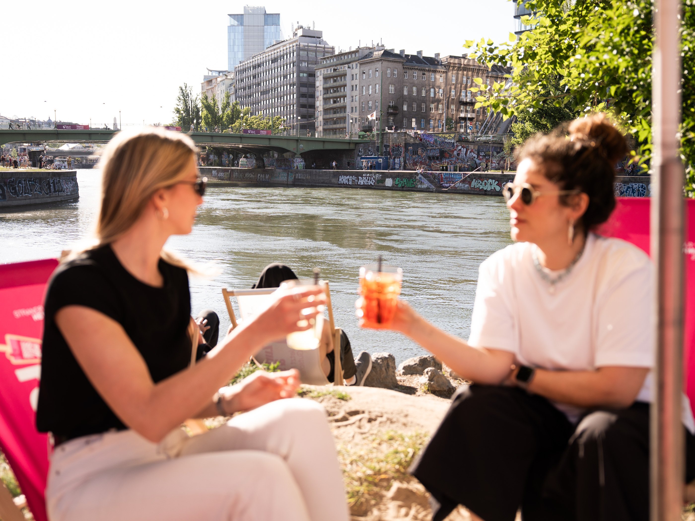 Due donne siedono allo Strandbar Herrmann sul Canale del Danubio, a Vienna, con un cocktail in mano. Sullo sfondo si intravedono un ponte e dei graffiti.