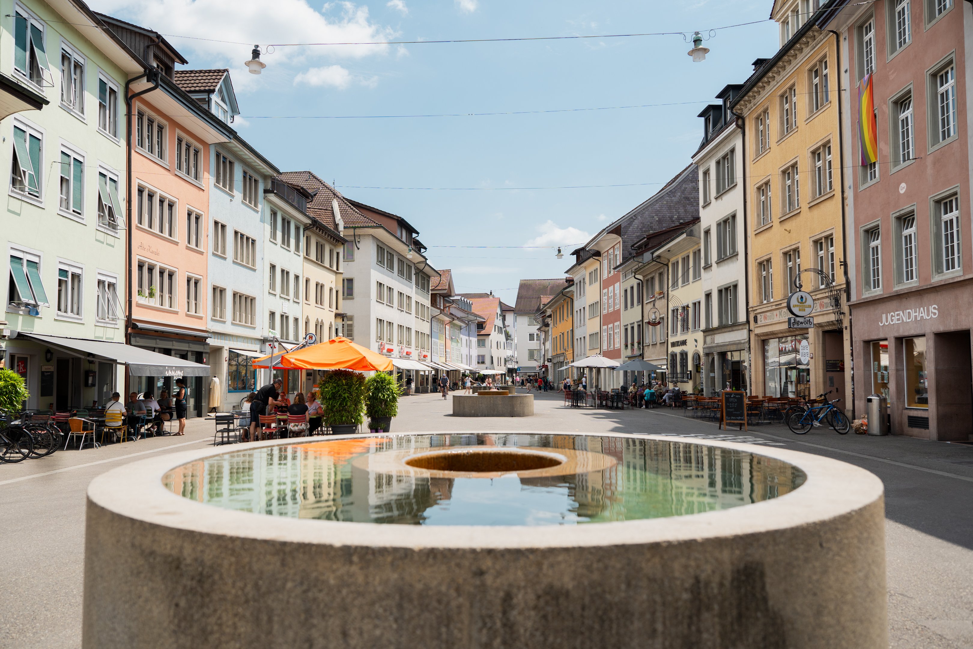 Une fontaine ronde avec un trou au centre se trouve dans une zone piétonne. Elle est bordée de façades multicolores.