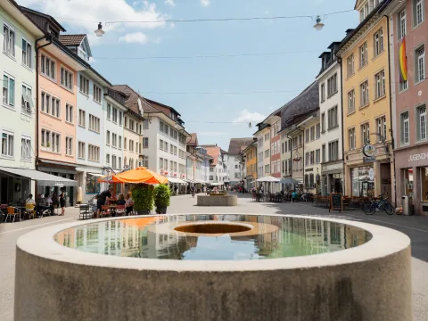 Une fontaine ronde avec un trou au centre se trouve dans une zone piétonne. Elle est bordée de façades multicolores.