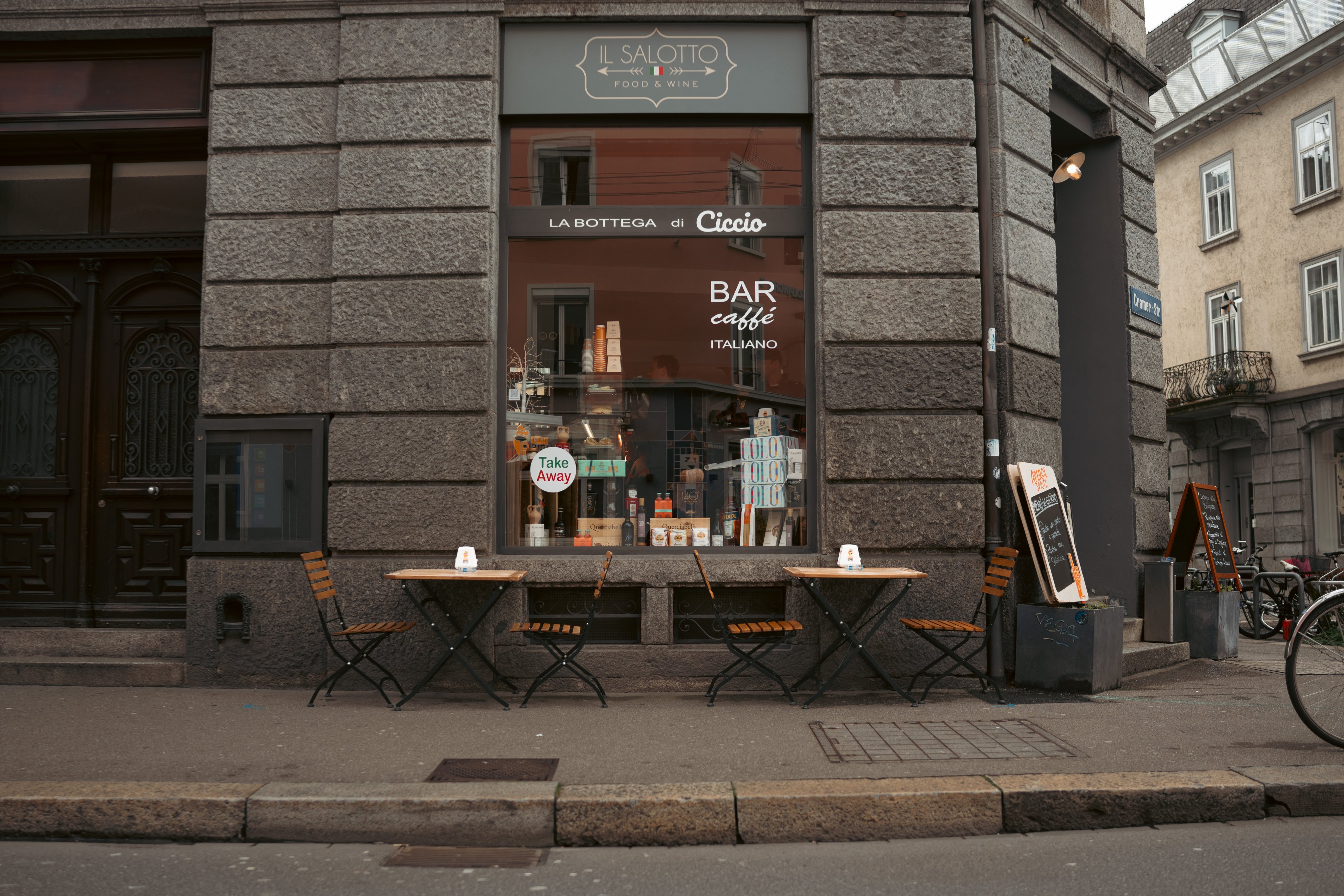 Des tables et chaises en bois se trouvent sur le chemin, devant un restaurant. Le restaurant est construit en pierres gris foncé. 