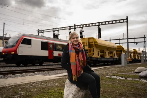Sandra Schwander vor einem Gleisfeld. Im Hintergrund fährt ein Zug der SBB. Der Zug ist ein Einheitswagen IV. Zudem sind drei gelbe Infrastrukturfahr