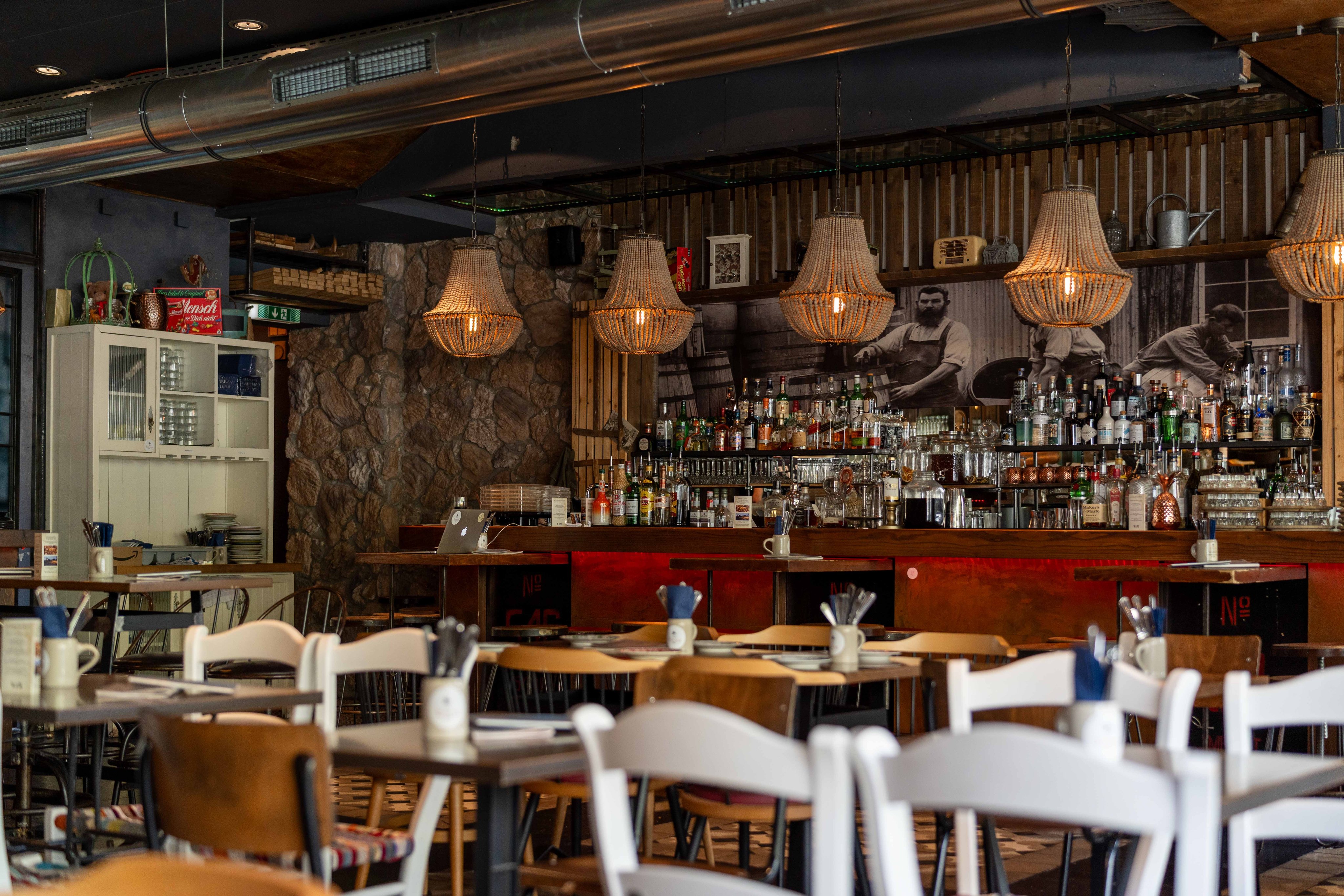 L’intérieur du restaurant: un comptoir avec toutes sortes de bouteilles et de verres sur les étagères. Ce comptoir est en pierre d’un côté et en bois de l’autre. Au premier plan, des tables en bois et des chaises. Au plafond, les tuyaux sont visibles et les luminaires ont des abat-jour tressés.