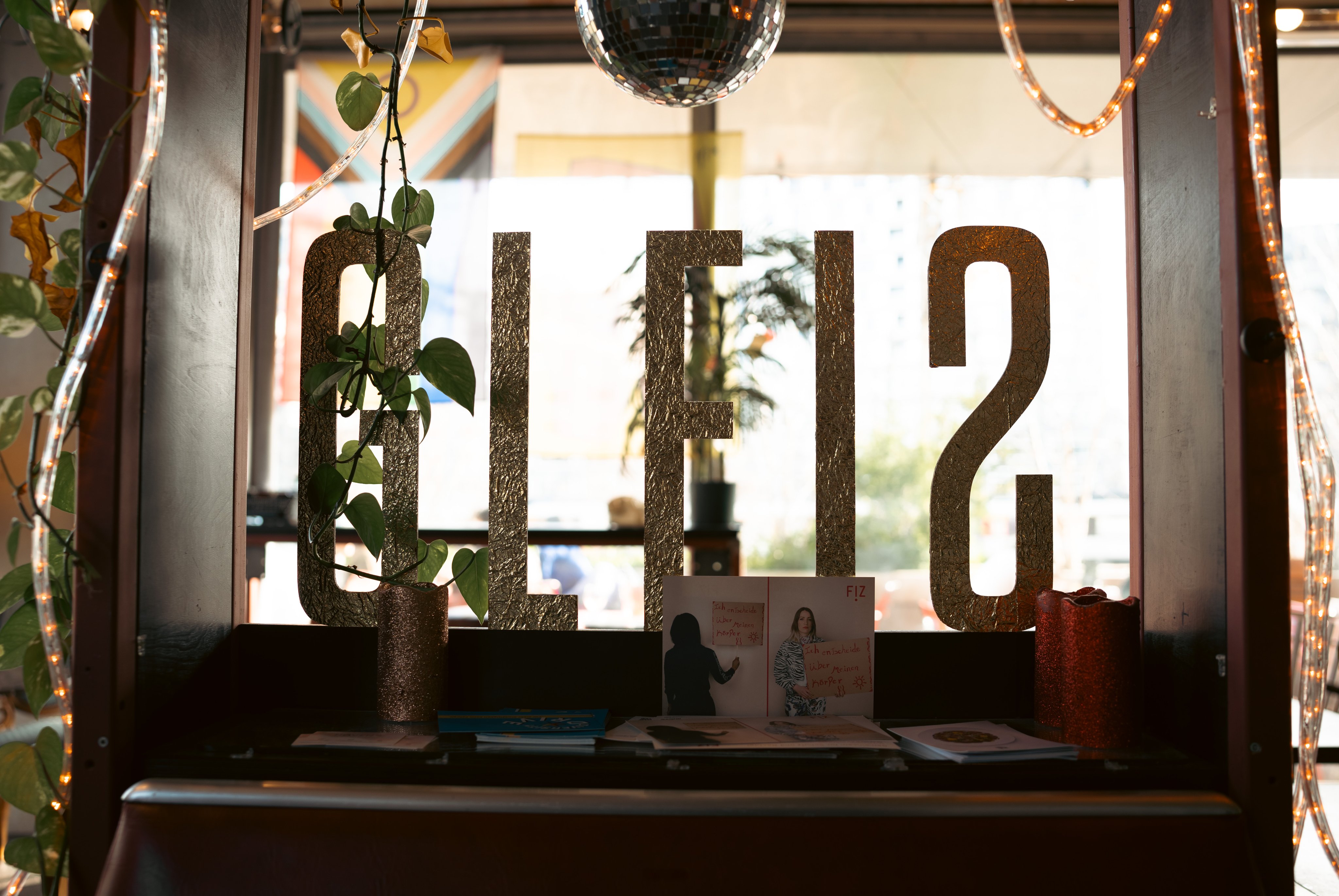 Dans le bar, devant une fenêtre, le nom Gleis est écrit en lettres dorées, entouré de bougies et de brochures sur le rebord de la fenêtre, de guirlandes lumineuses et de plantes suspendues au plafond.