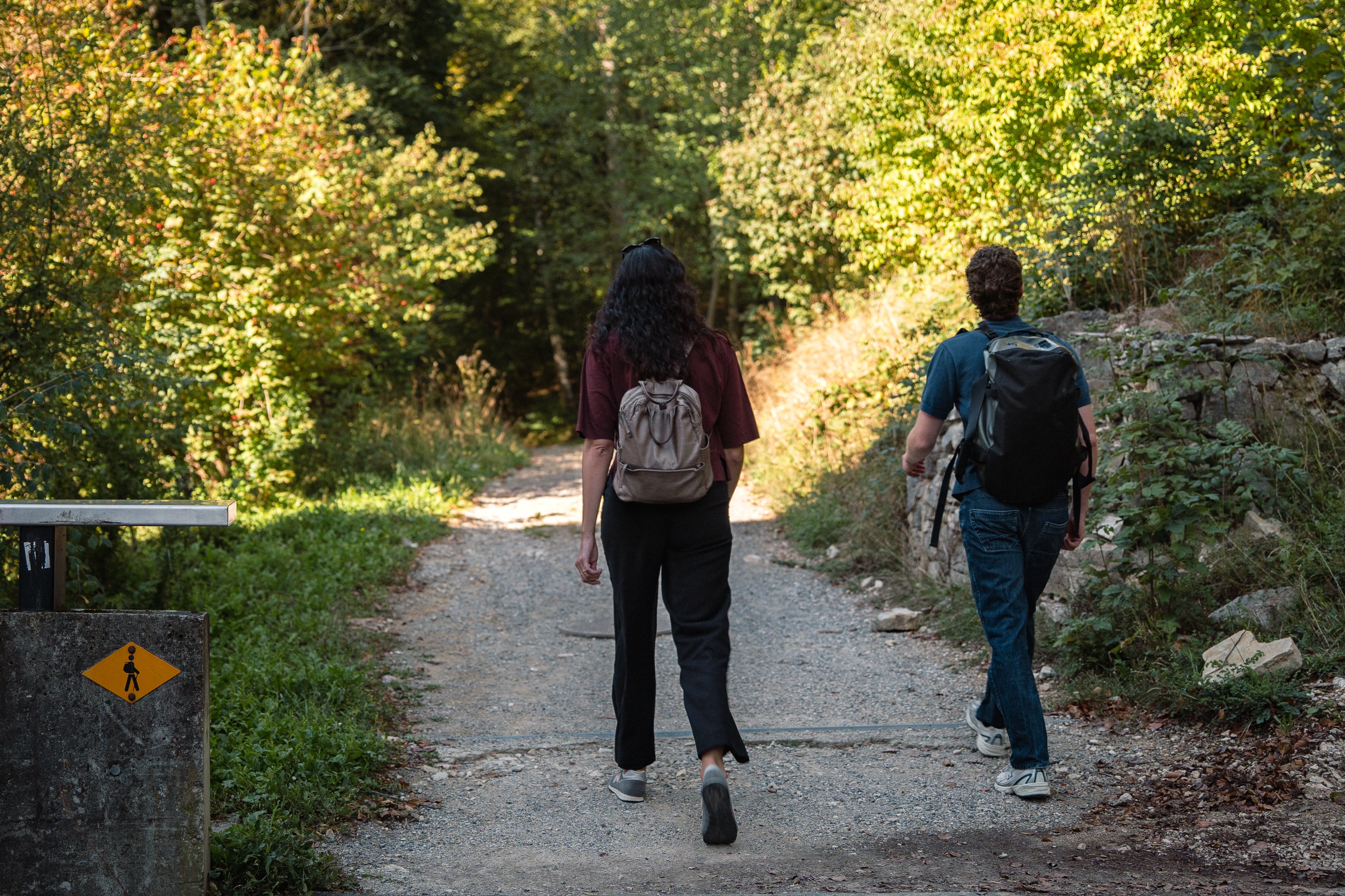 Due persone camminano lungo un sentiero sterrato in direzione del bosco. In basso a sinistra si vede un cartello escursionistico giallo fissato a un muro.