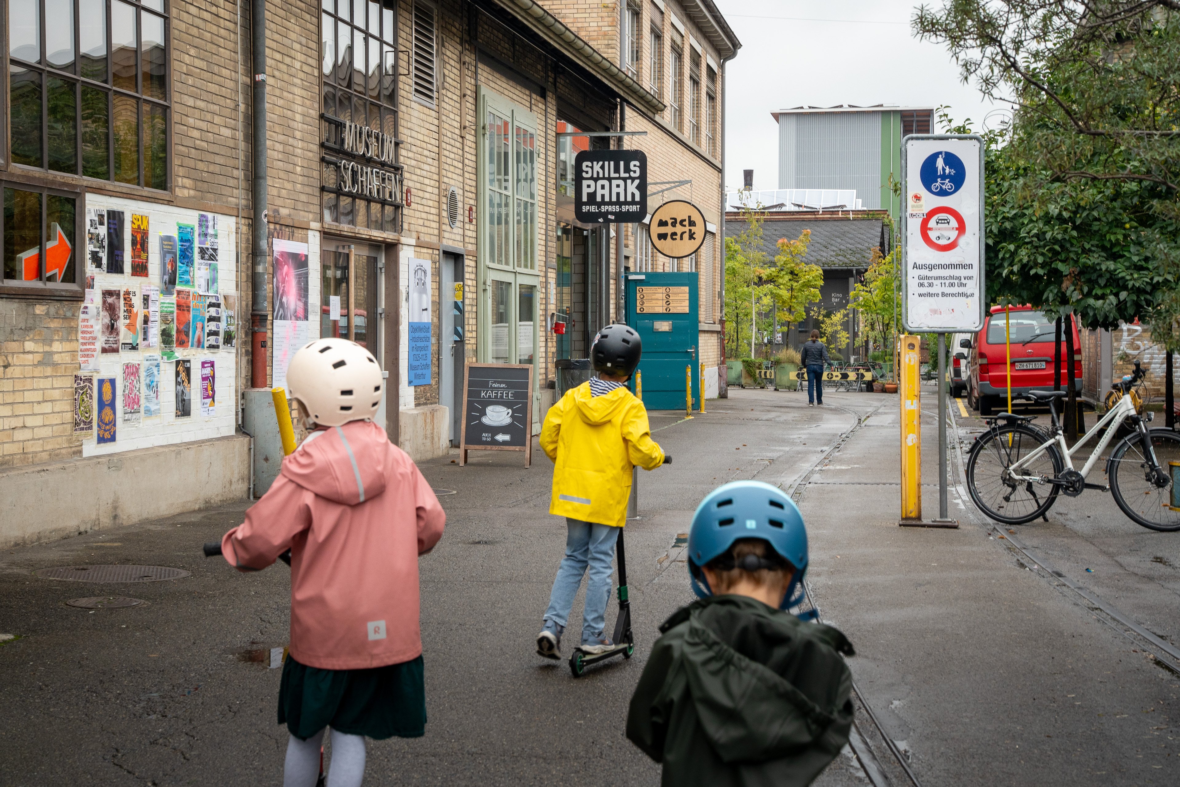 Drei Kinder auf dem Trottinett bei Regenwetter. 