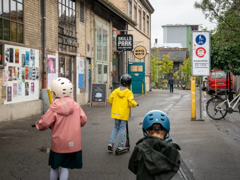Drei Kinder auf dem Trottinett bei Regenwetter.