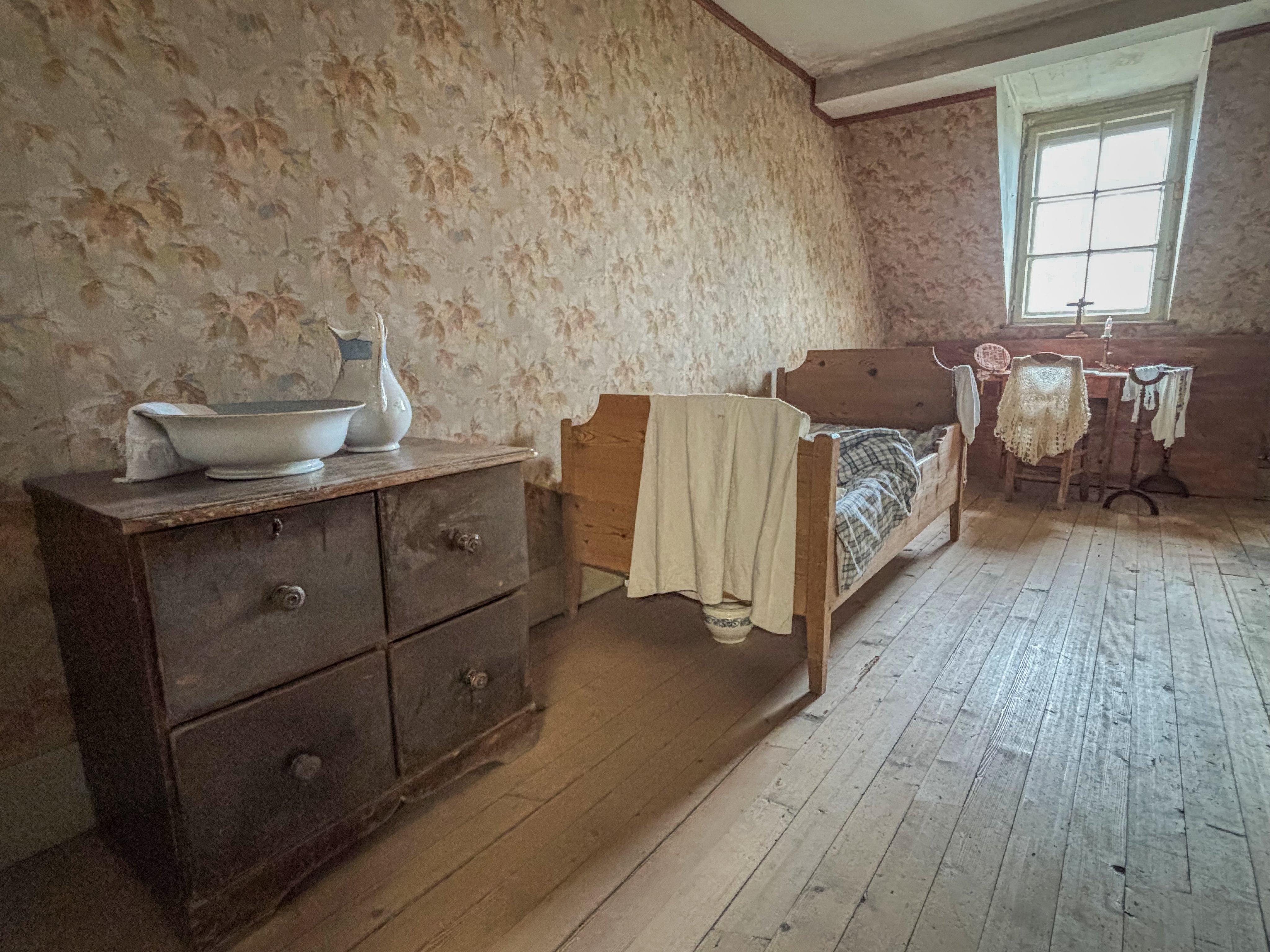 In einem Zimmer mit Holzboden steht eine Kommode mit einer Waschschüssel, ein Bett sowie ein Tisch mit einem Stuhl davor.