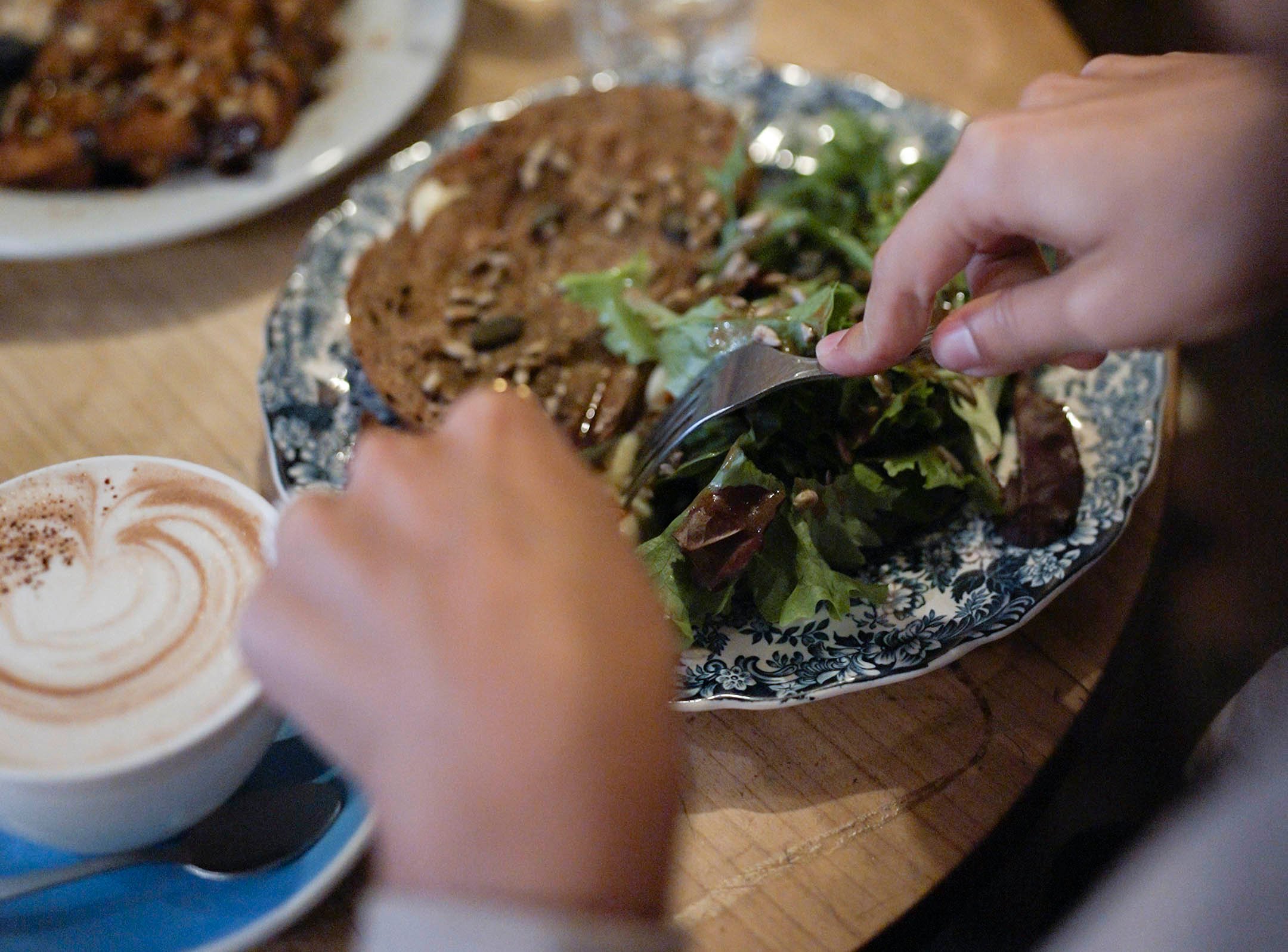 Main préparant une salade à côté d’un toast au fromage et aux tomates séchées joliment présenté et d’une tasse de cappuccino surmontée de mousse en forme de cœur.