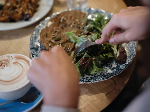 Main préparant une salade à côté d’un toast au fromage et aux tomates séchées joliment présenté et d’une tasse de cappuccino surmontée de mousse en forme de cœur.