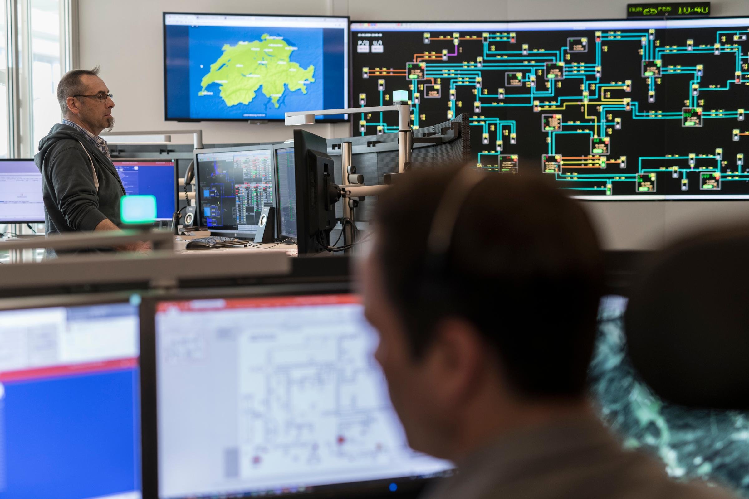 Im Vordergrund schaut ein Mann auf viele Bildschirme, dieser Teil des Fotos ist leicht unscharf. Im Hintergrund schaut ein Mann mit Bart ebenfalls auf viele Bildschirme. An der Wand des Raums sind grosse Bildschirme ersichtlich welche das Wetter zeigen und die Kraftwerke abbilden.