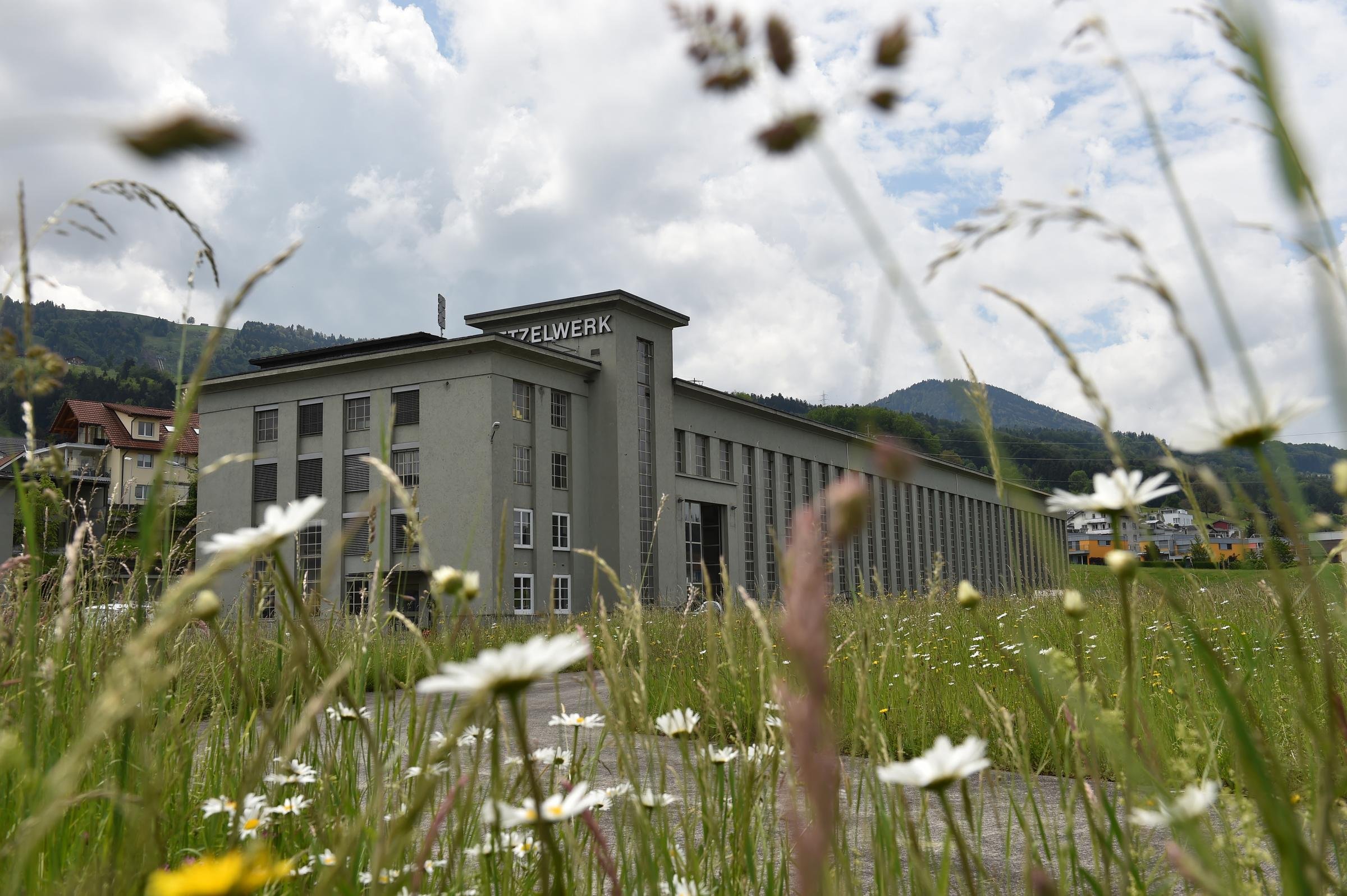 Ein grosses graues Gebäude mit langen Fenstern. Das Gebäude wirkt alt. Es ist mit einem Schriftzug «Etzelwerk» beschriftet. Im Vordergrund ist eine Wiese sowie Stromleitungen. 