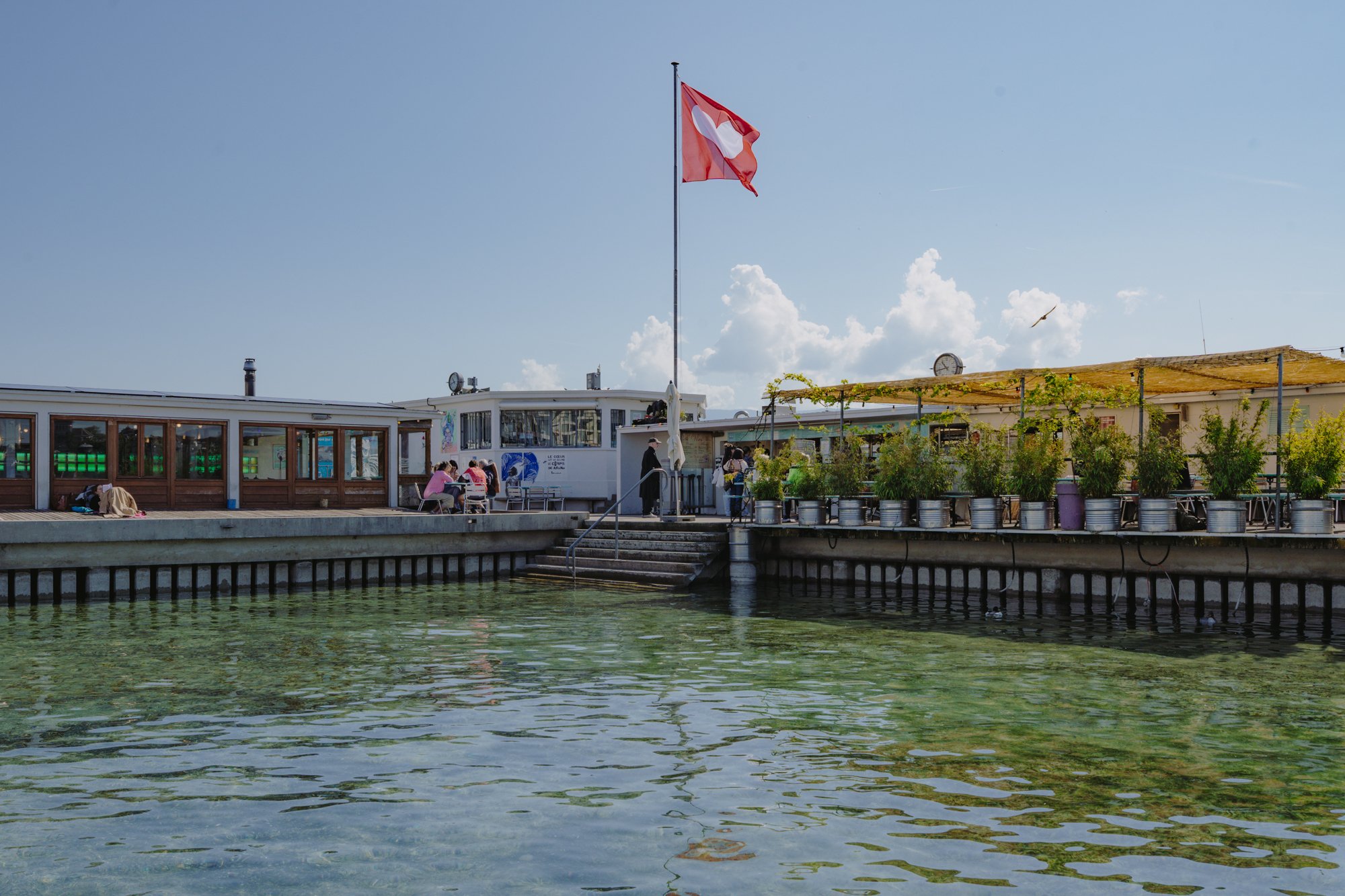 Les Bains des Pâquis à Genève avec drapeau suisse, terrasse ensoleillée, jardinières et eau claire.