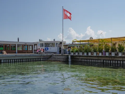 Les Bains des Pâquis à Genève avec drapeau suisse, terrasse ensoleillée, jardinières et eau claire.