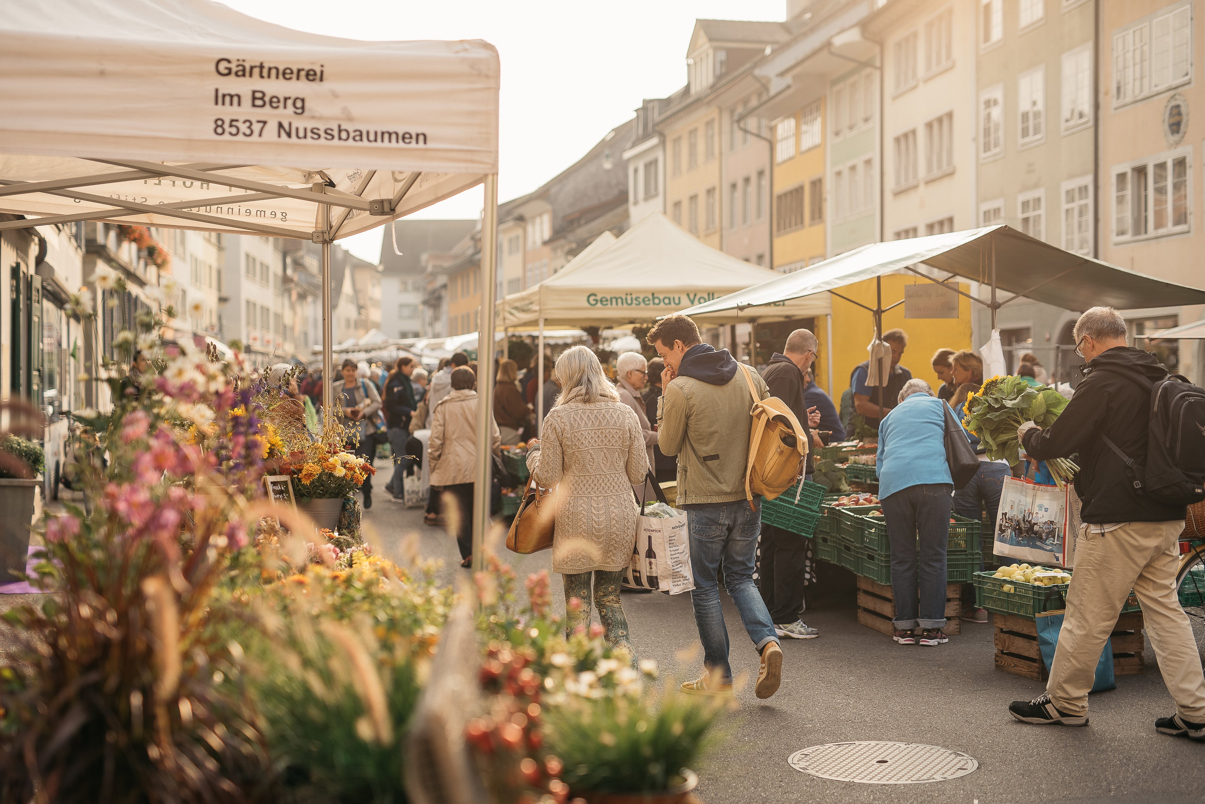 Des gens passent devant des étals avec des fleurs et des légumes, devant les façades des maisons de la vieille ville.
