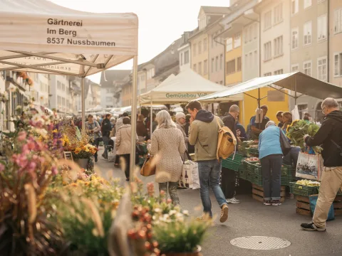 Des gens passent devant des étals avec des fleurs et des légumes, devant les façades des maisons de la vieille ville.