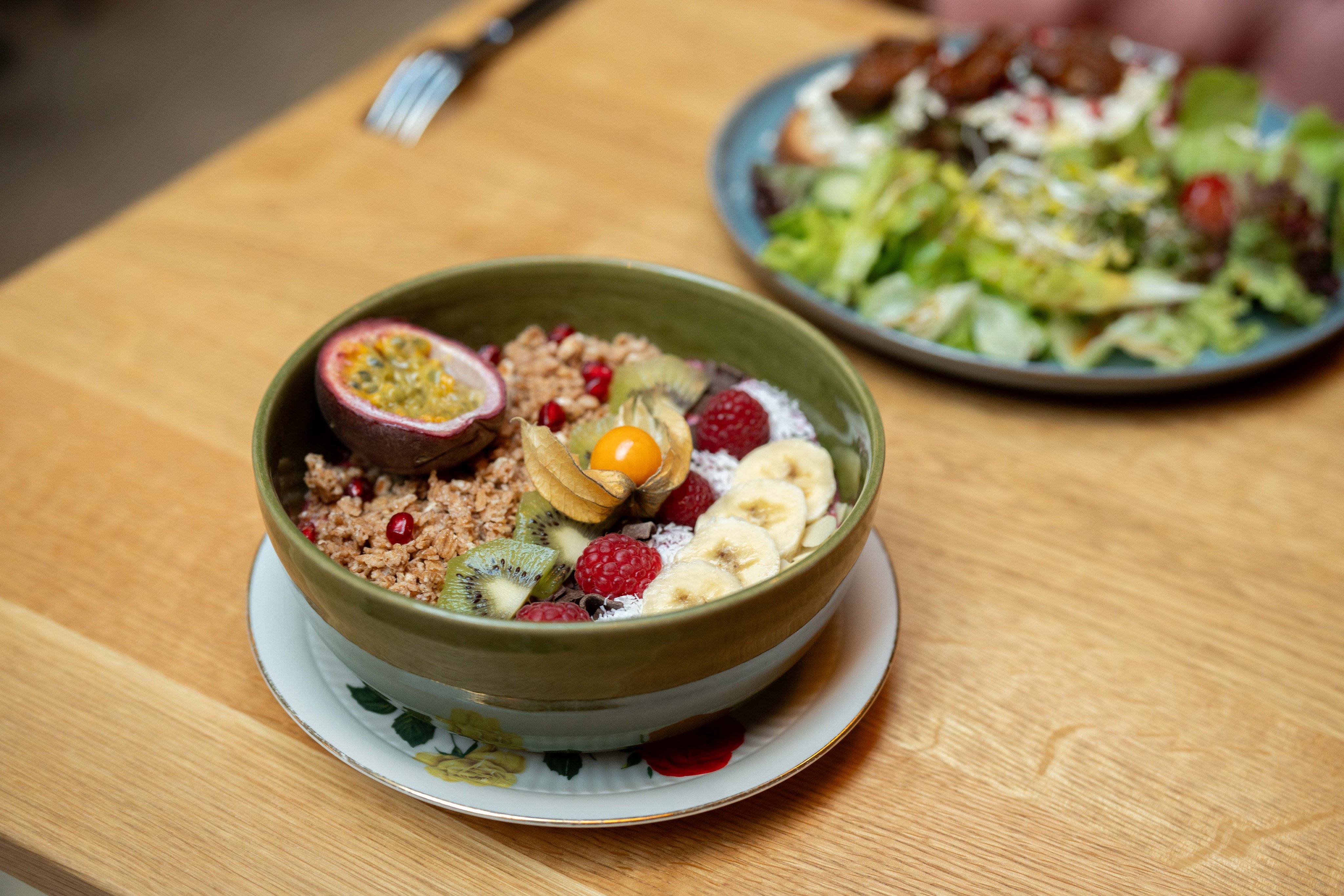 Muesli, fruits et noix dans une assiette verte posée sur une table en bois, avec une salade à l’arrière-plan.